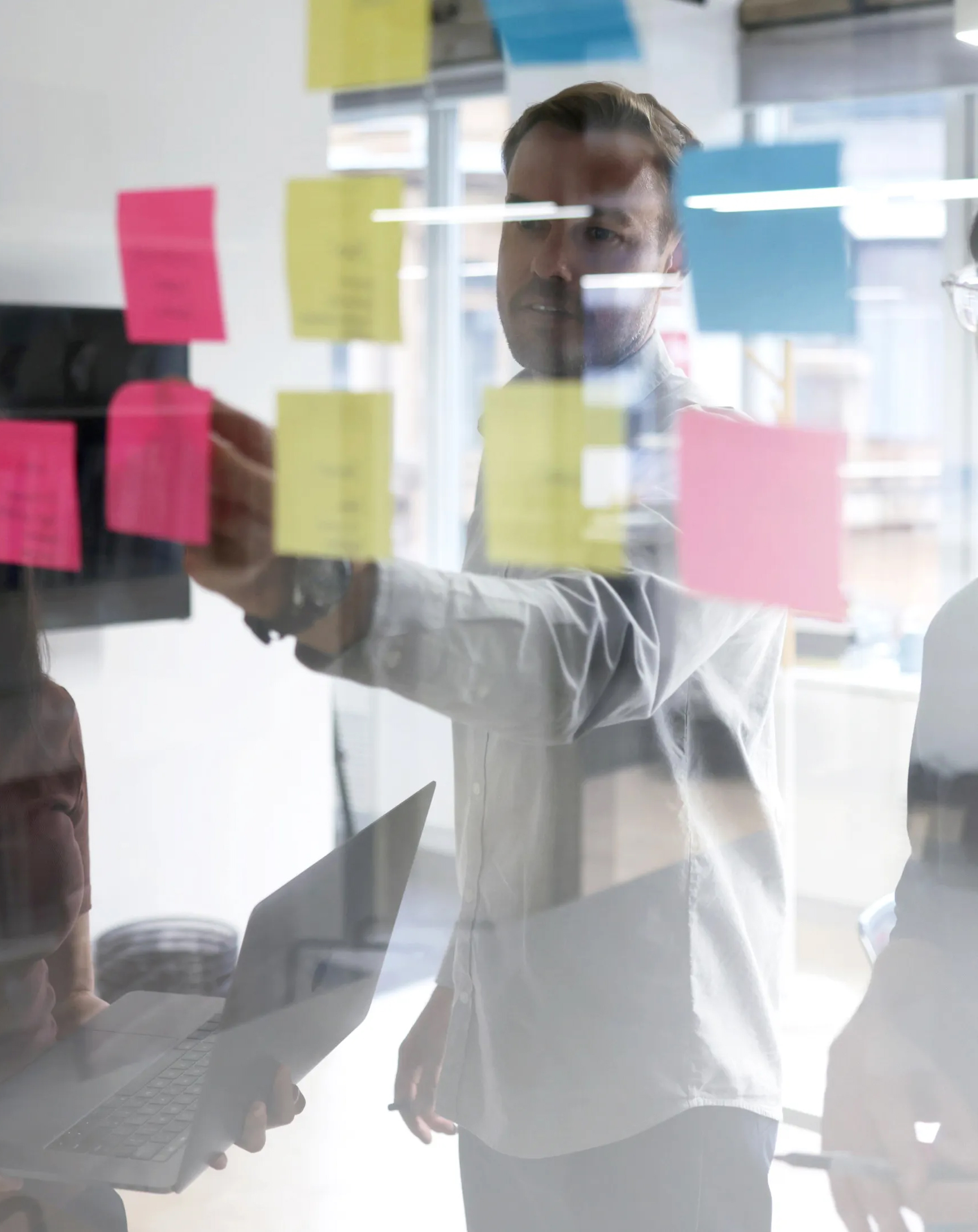 Man pointing at colorful sticky notes on glass during a team brainstorming session in an office.
