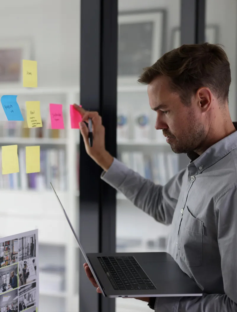 Man holding laptop and writing on pink sticky note on glass wall with other colorful notes.
