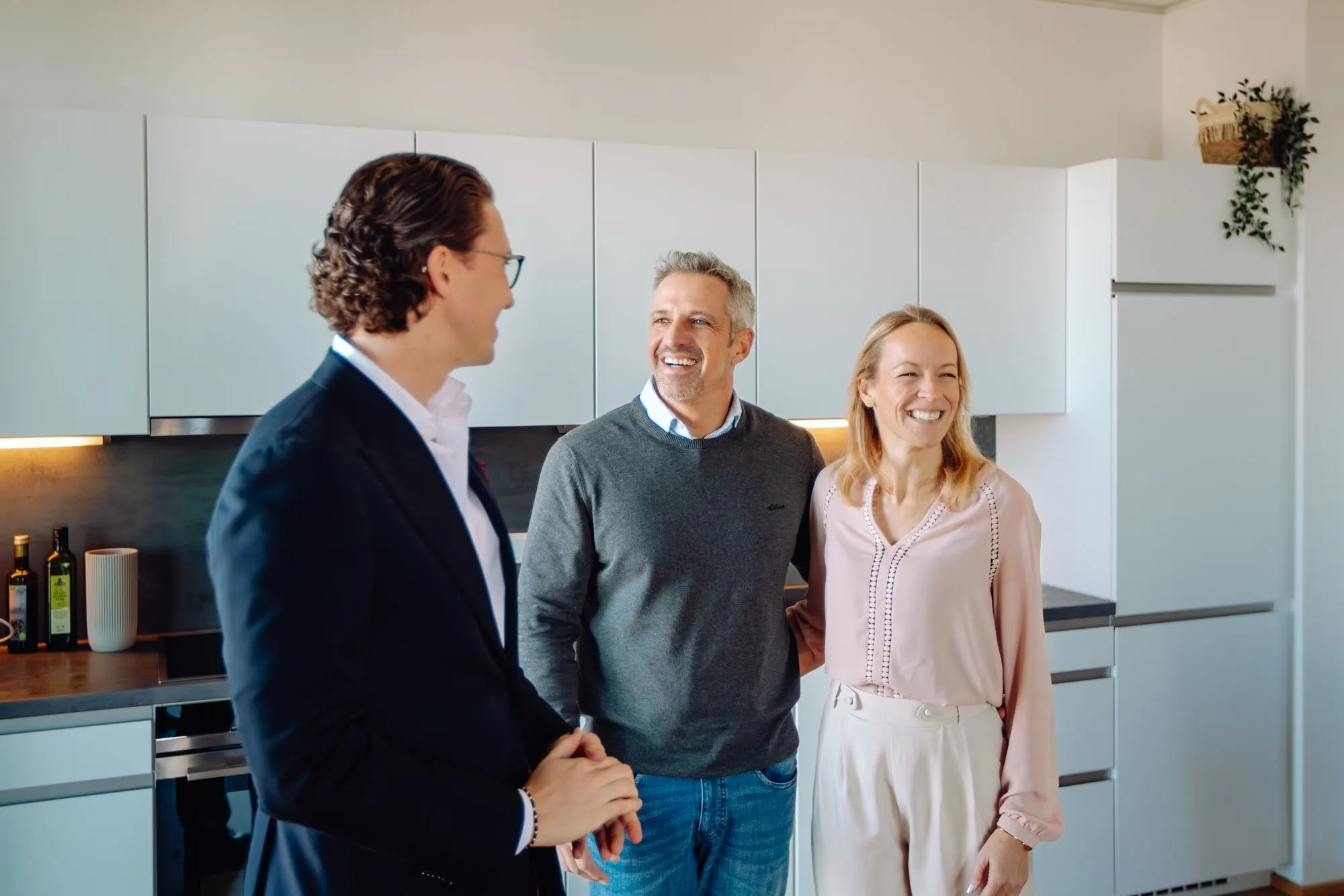 A couple smiling and standing in a modern kitchen with a man in a suit talking to them.