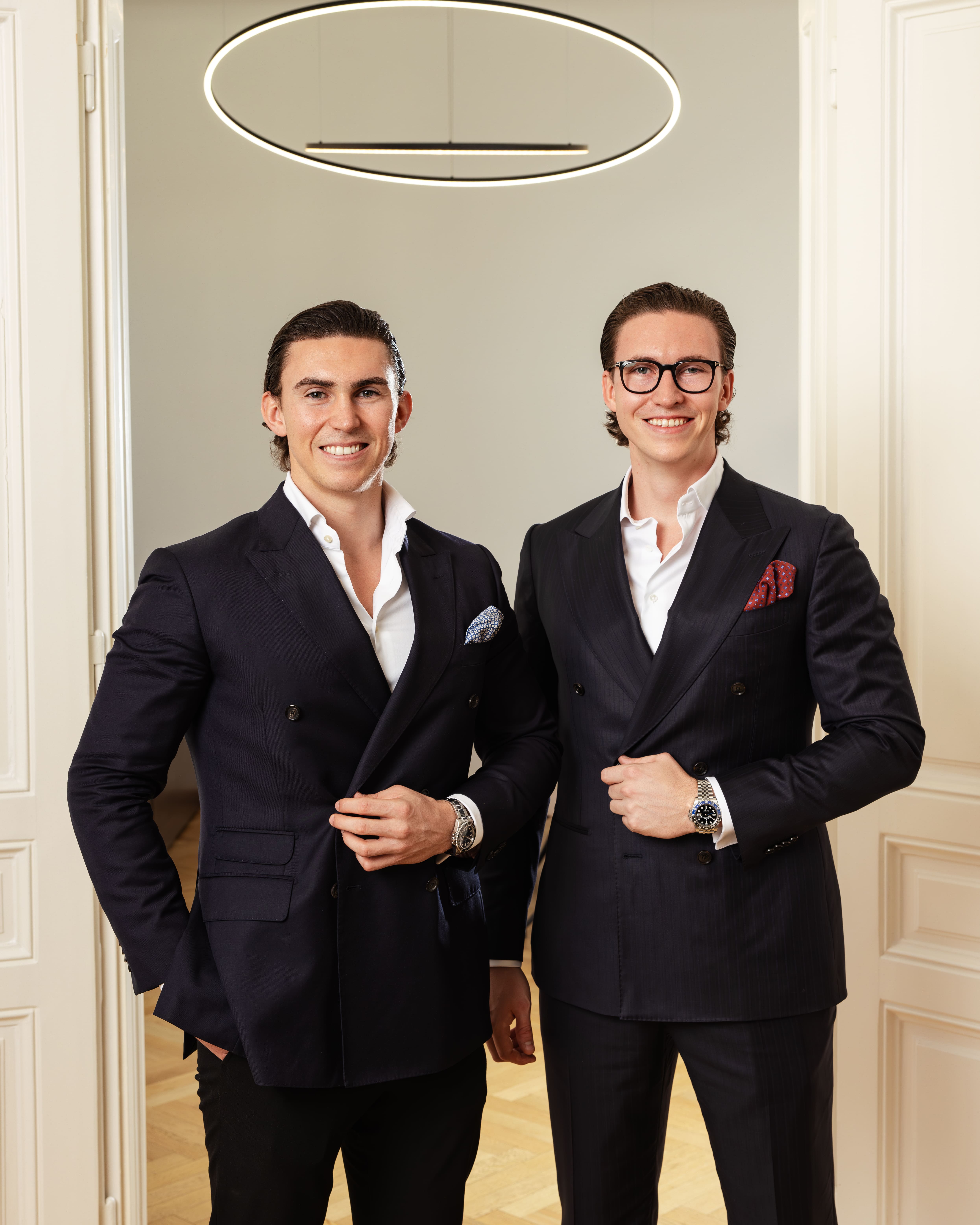 Man in blue shirt shaking hands with man in black suit while a woman in white sweater smiles beside them in a living room.