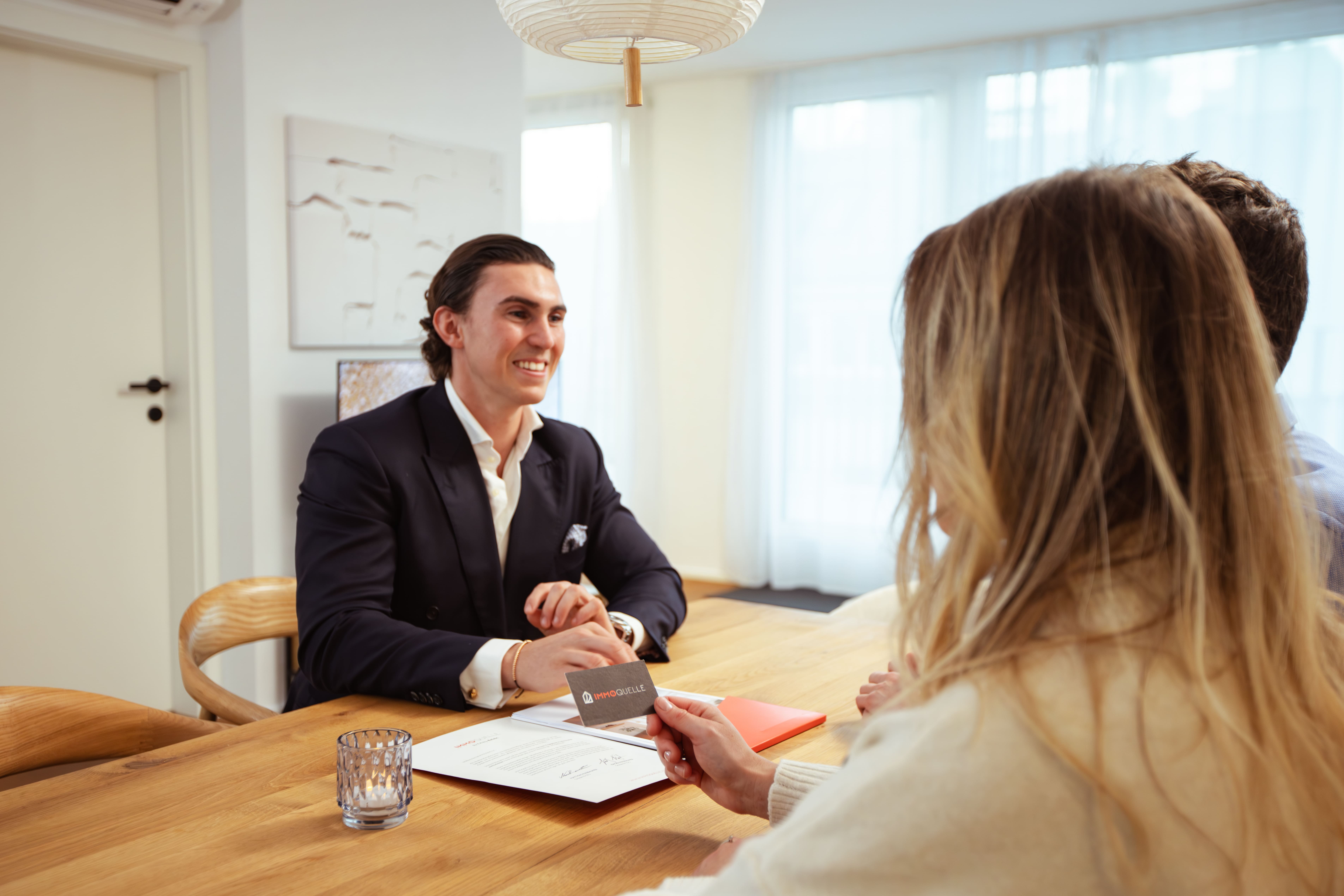 A couple dressed in fall clothing talking and smiling with a man in a black suit near a building and lawn.