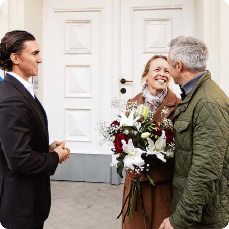 Smiling woman holding a bouquet of flowers is greeted by a man in a green jacket while another man in a suit watches.