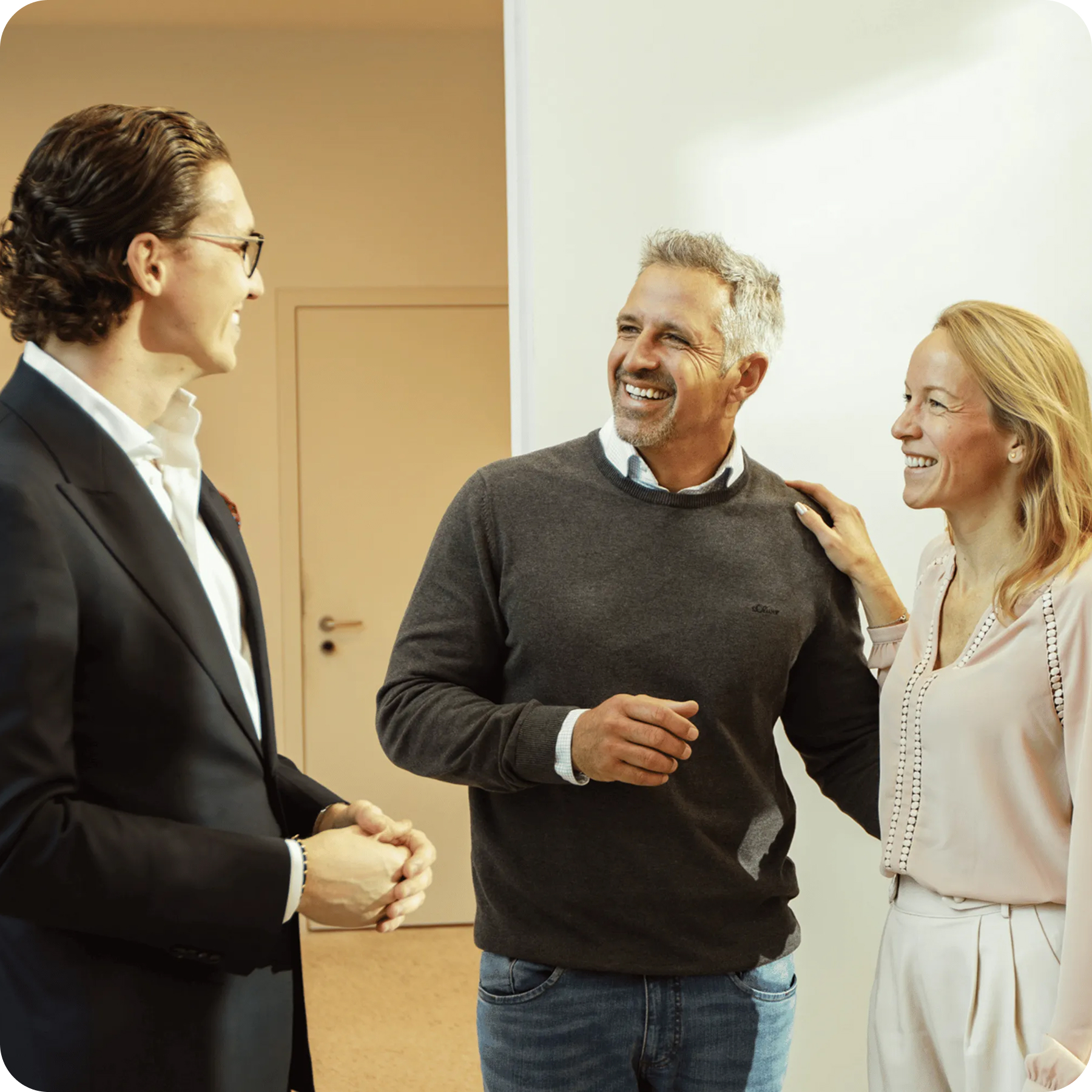 Two men shaking hands and smiling while a woman stands between them with a cheerful expression indoors.