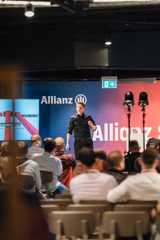 Ein Vortragender steht auf einer Bühne vor einem Publikum, im Hintergrund sind Banner mit „Allianz“ und weiteren Schriftzügen zu sehen.