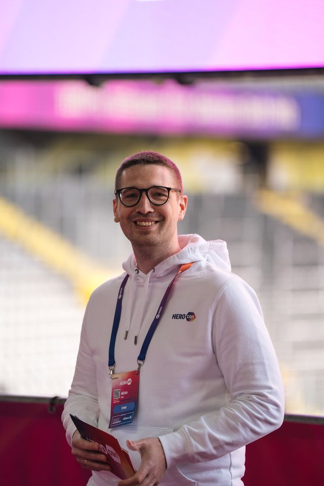 Person im weißen HEROcon-Hoodie mit Event-Lanyard und Namensschild, hält Karten in der Hand, im Hintergrund Stadion-Atmosphäre.