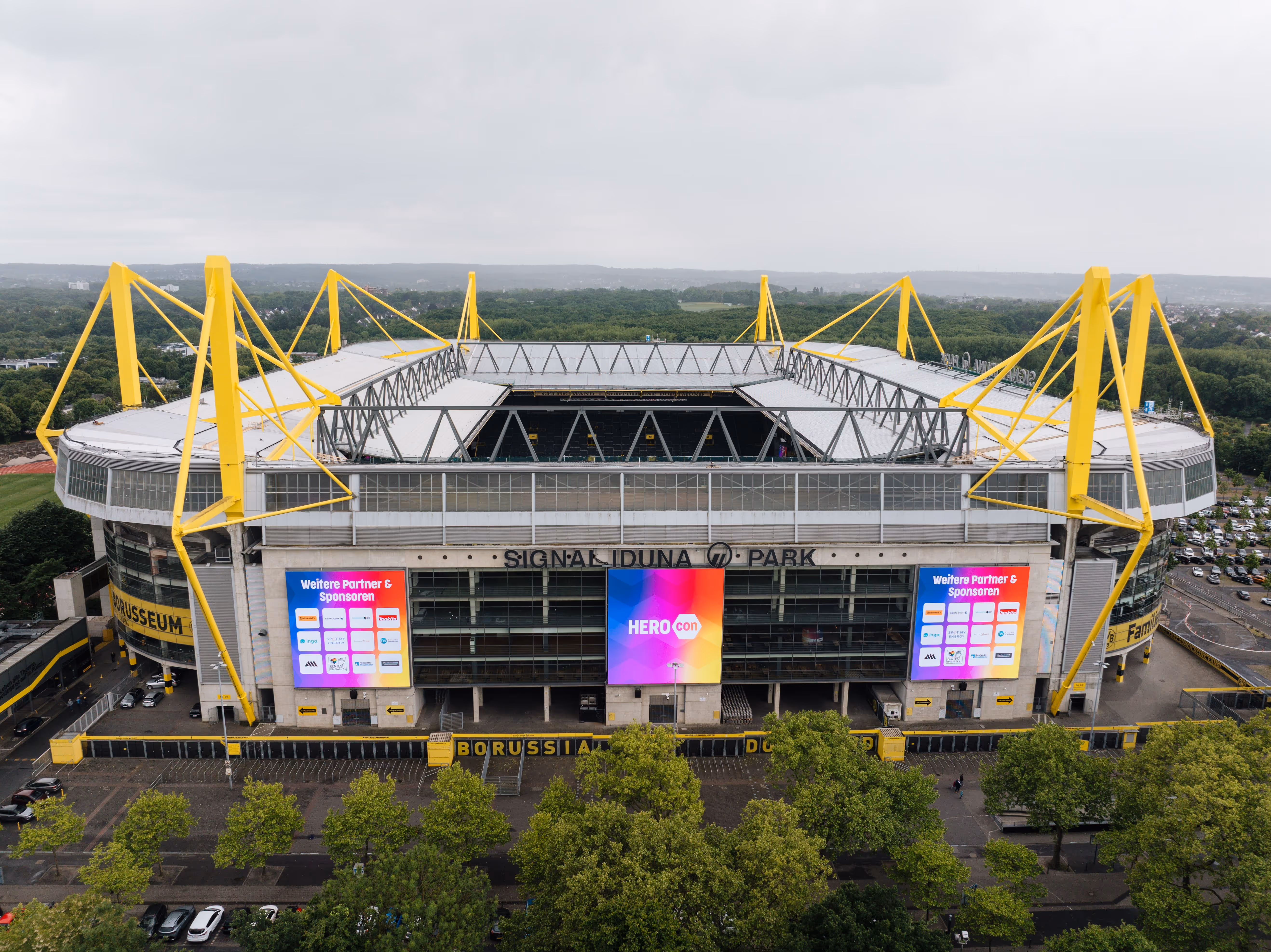 Das Fußballstadion Signal Iduna Park in Dortmund, mit gelben Stahlträgern und großen LED-Bildschirmen an der Fassade, umgeben von Bäumen und Parkplätzen.