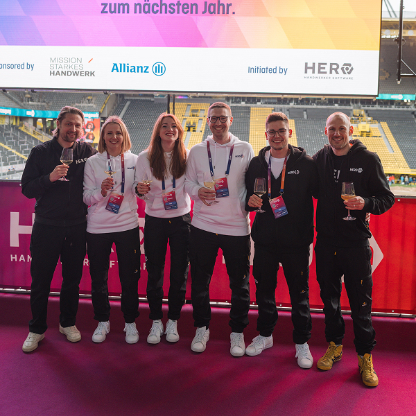 Sechs Personen mit Konferenz-Badges und Getränken posieren gemeinsam vor einer HEROcon-Bühne im Stadion, im Hintergrund sind Tribünen zu sehen.