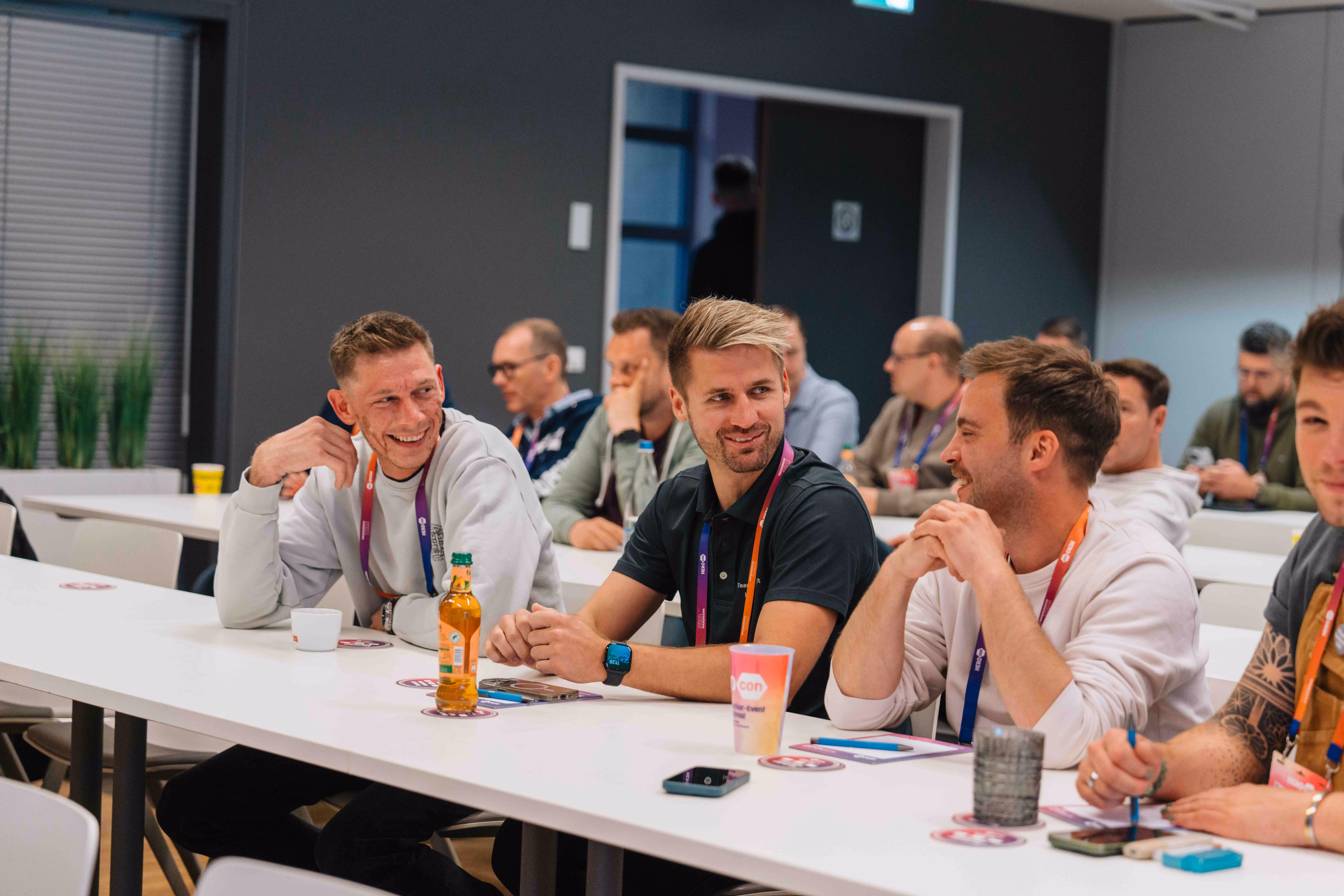 Mehrere Personen sitzen in einem hellen Seminarraum an langen Tischen und hören aufmerksam zu.