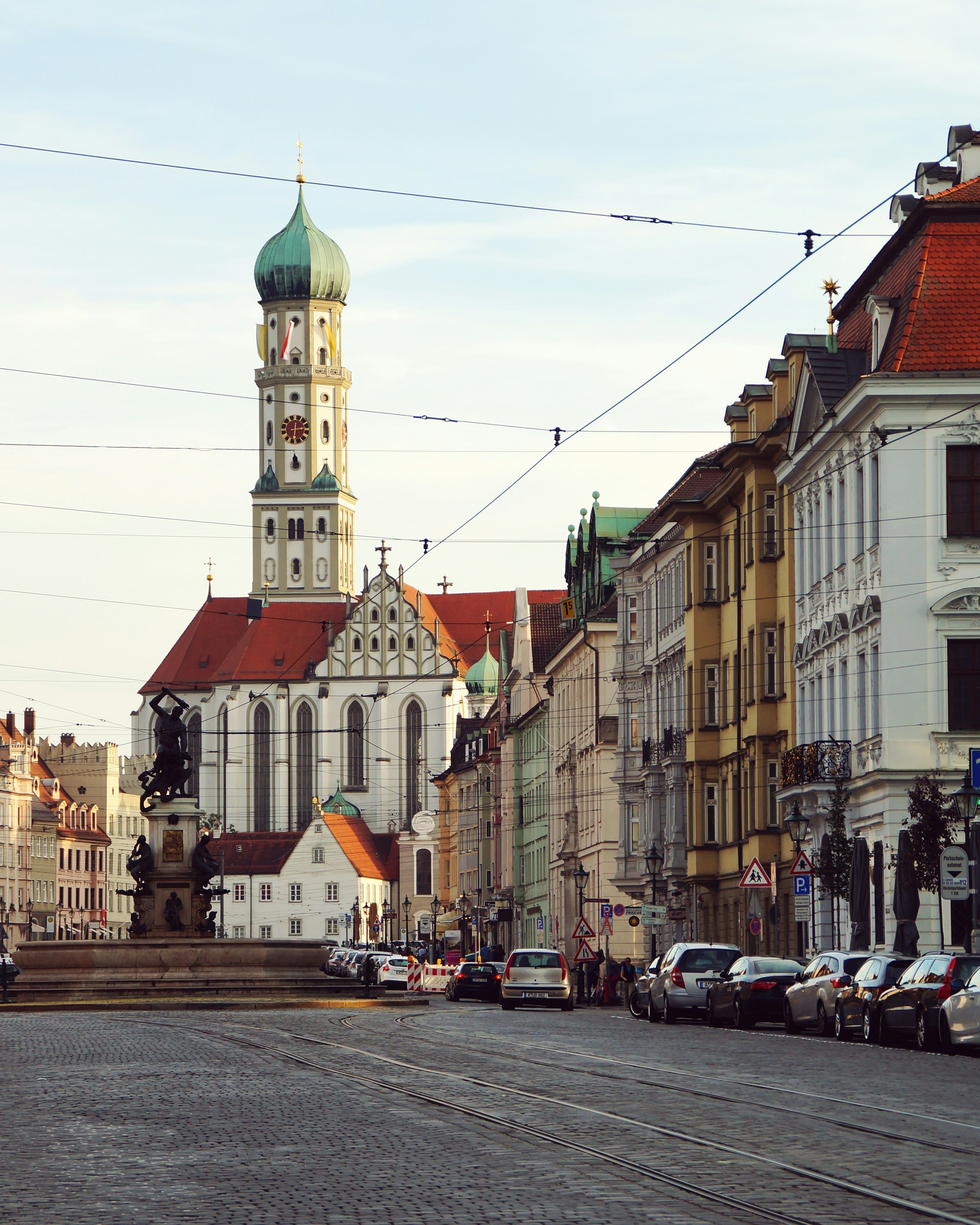 Blick auf eine Straße mit Kopfsteinpflaster, geparkten Autos und historischen Gebäuden, dominiert von einem großen Kirchturm mit grünem kupfernen Dach.