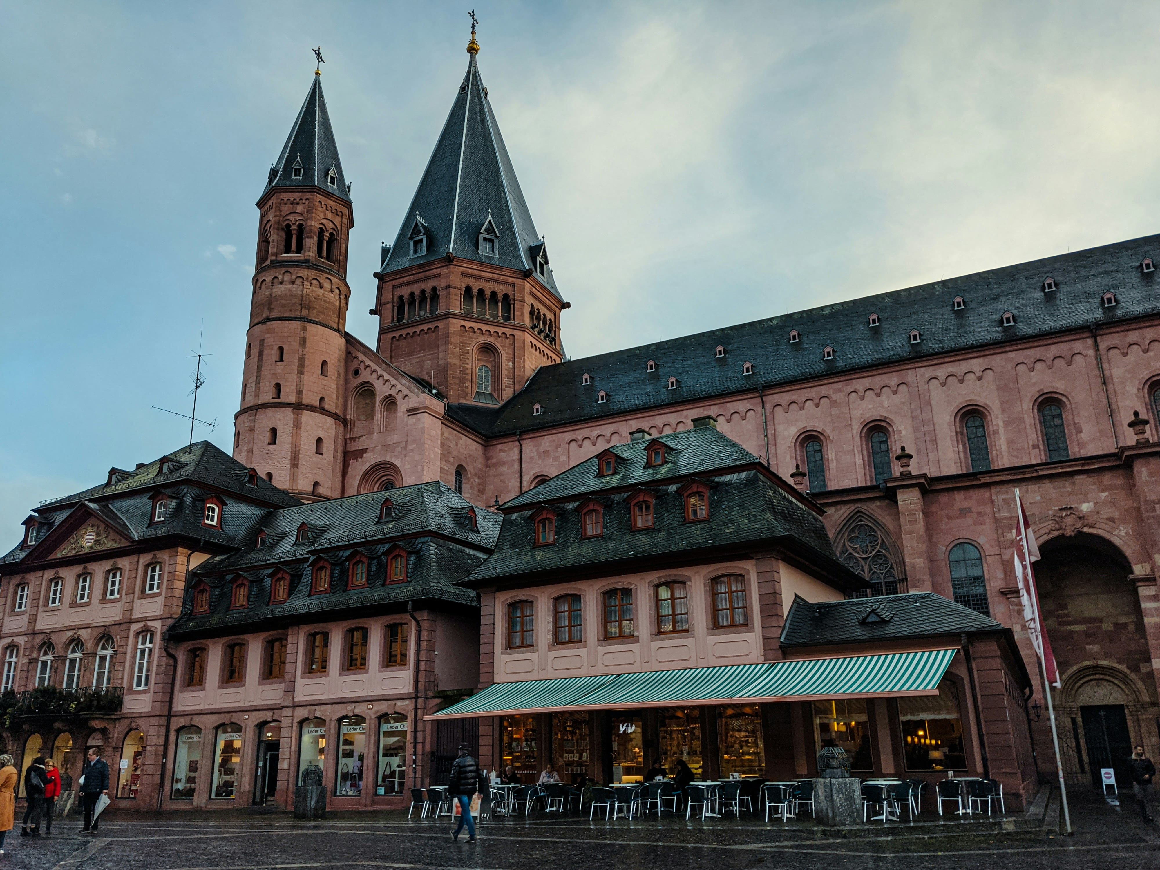 Historische Gebäude mit steinernen Türmen und einem Café mit grün-weiß gestreiftem Sonnenschutz in einem belebten Platz.
