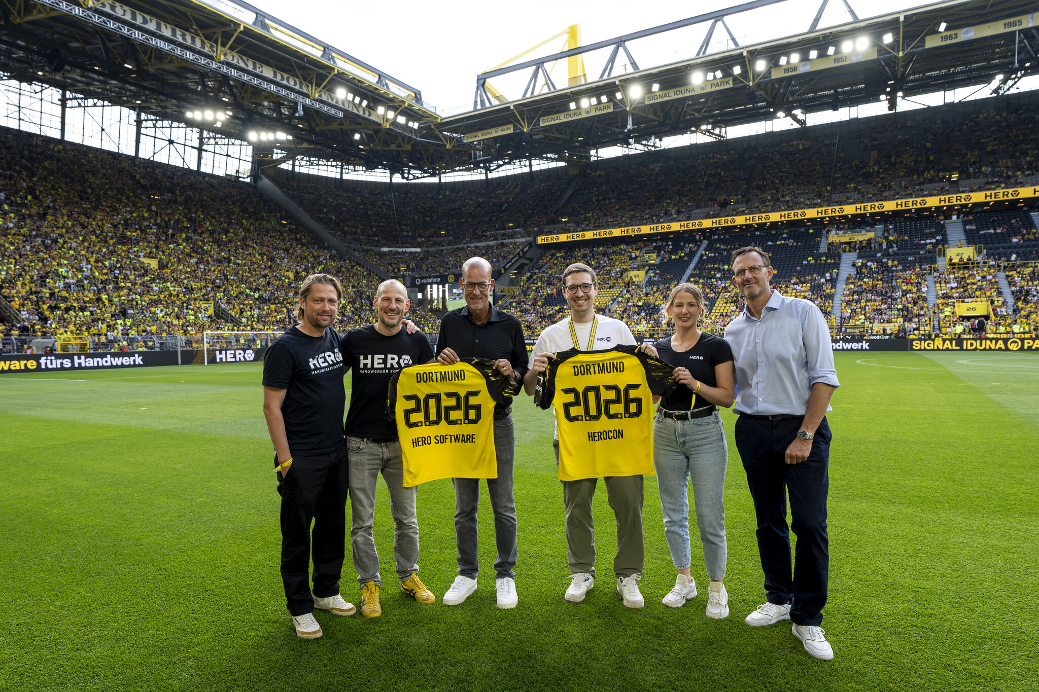 Sechs Personen stehen im Fußballstadion Signal Iduna Park auf dem Spielfeld, zwei halten gelbe Trikots mit der Aufschrift Dortmund 2026, umgeben von Zuschauern auf den Rängen.