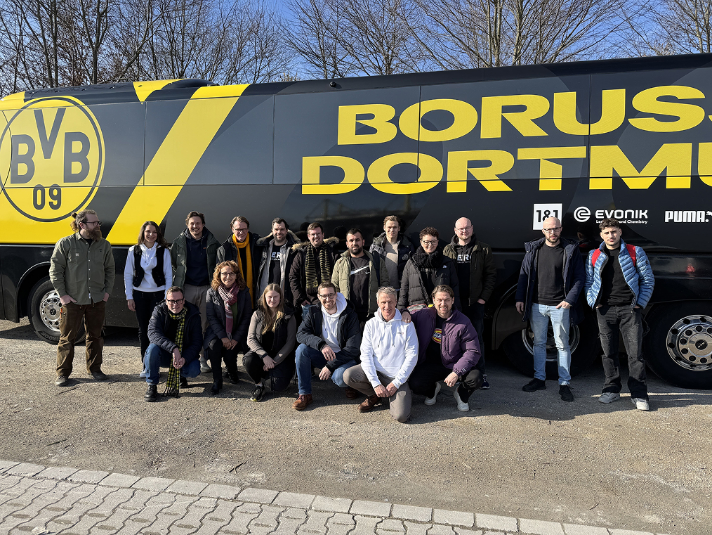Eine Gruppe von 17 Personen posiert vor einem schwarzen und gelben Borussia Dortmund Mannschaftsbus an einem sonnigen Tag.