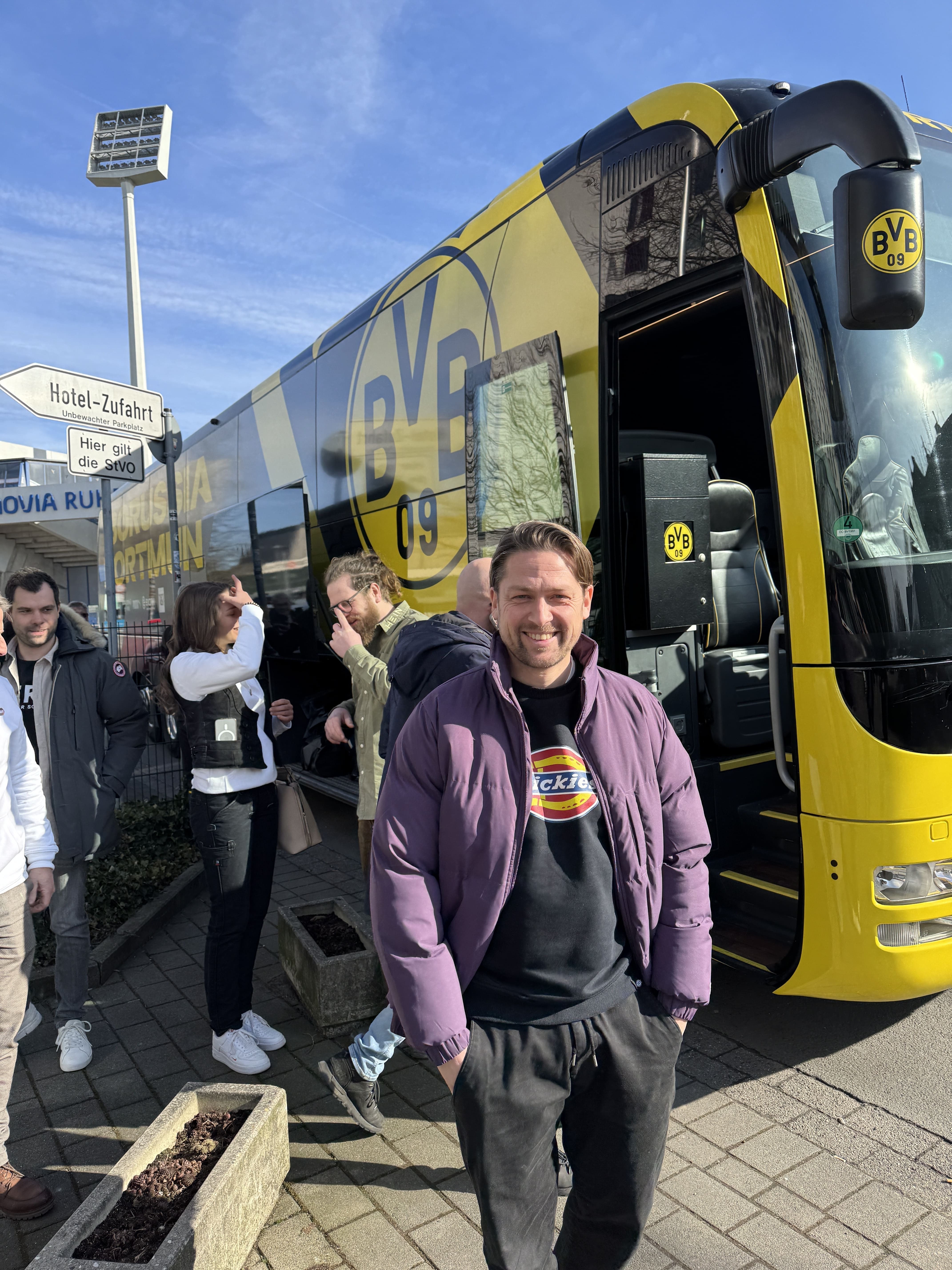 Mann in lila Jacke steht lächelnd vor einem gelb-schwarzen BVB 09 Mannschaftsbus, im Hintergrund weitere Personen und ein Schild mit der Aufschrift Hotel-Zufahrt.