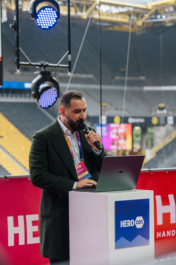 Ein Mann mit Bart spricht in ein Mikrofon, während er vor einem Laptop auf einem Podium bei einer Veranstaltung mit der Aufschrift HEROcon steht.