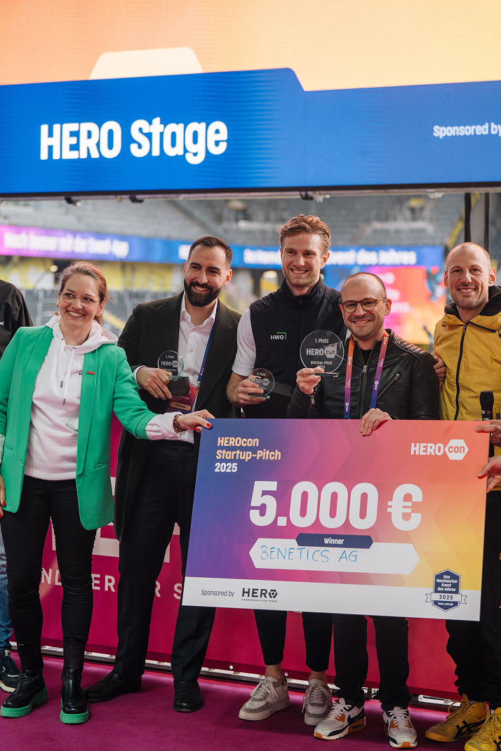 Gruppe von Menschen auf der HERO Stage hält einen großen Scheck über 5.000 Euro für Gewinner Benetics AG beim HEROCON Startup-Pitch 2025.