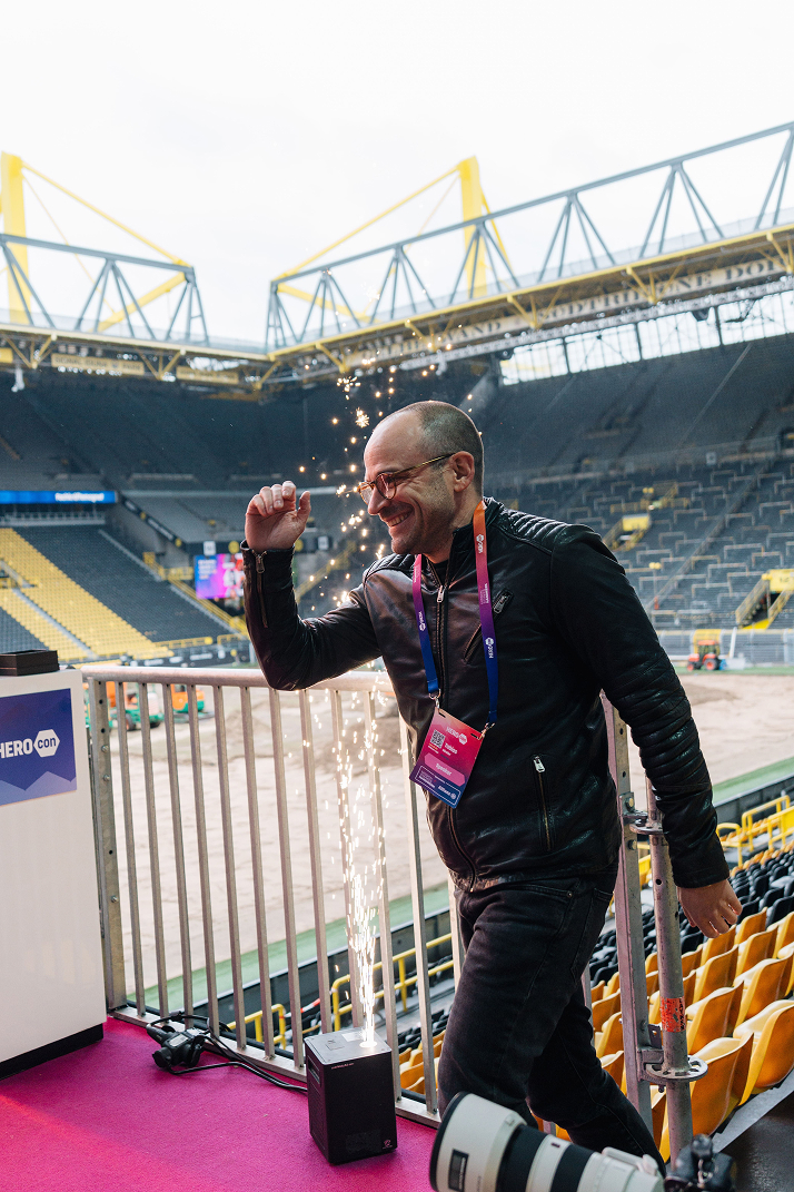 Mann in schwarzer Lederjacke und Brille bewegt sich lächelnd neben einem Feuerwerksgerät in einem großen Stadion mit gelben Sitzreihen.
