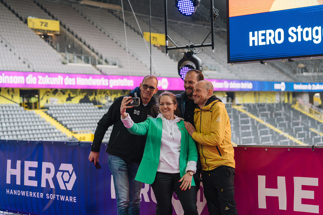 Vier Personen machen zusammen ein Selfie bei einer Veranstaltung in einem großen Stadion mit Hero Handwerker Software-Bannern im Hintergrund.