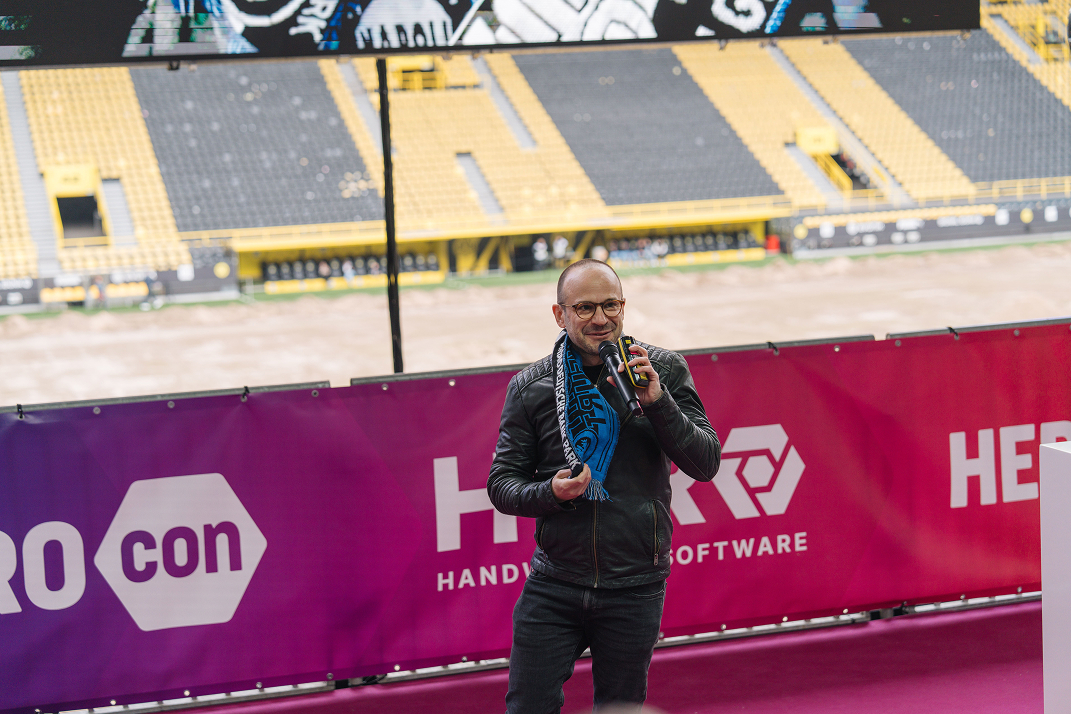 Ein Mann mit Brille und Lederjacke hält ein Mikrofon und spricht vor einer lila Werbebande in einem Stadion mit gelb-schwarzen Sitzen.
