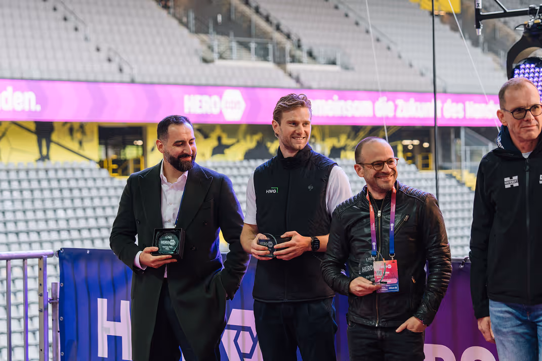 Drei Männer stehen nebeneinander in einem Stadion, jeder hält einen HEROcon-Preis, sie lächeln und tragen unterschiedliche Outfits.