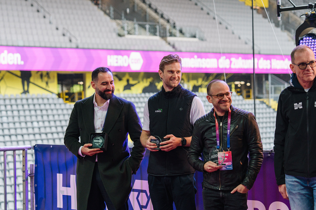 Drei Männer stehen nebeneinander in einem Stadion, jeder hält einen HEROcon-Preis, sie lächeln und tragen unterschiedliche Outfits.