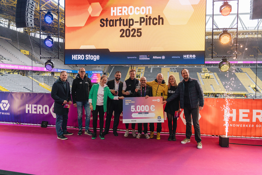 Gruppe von neun Personen steht auf einer Bühne mit einem großen Scheck über 5.000 Euro bei der HeroCon Startup-Pitch 2025 Veranstaltung in einem Stadion.