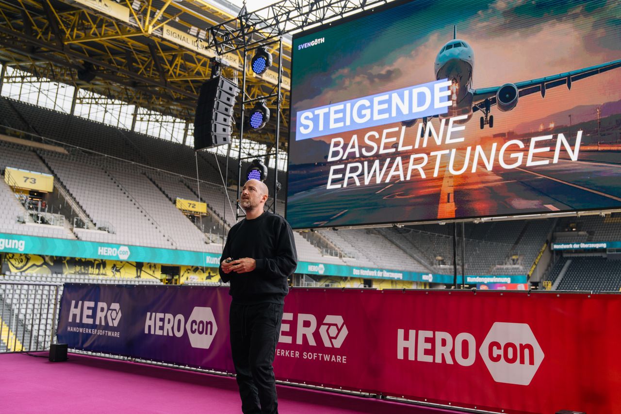 Mann in schwarzer Kleidung steht auf einer Bühne mit einem großen Bildschirm, der ein Flugzeug und den Text 'STEIGENDE BASELINE ERWARTUNGEN' zeigt, im Inneren eines großen Stadions.