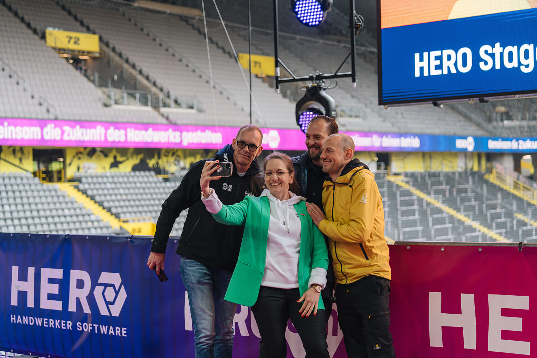 Vier Personen machen ein Selfie vor einer Bühne mit Bannern der HERO Handwerker Software in einer großen Halle.
