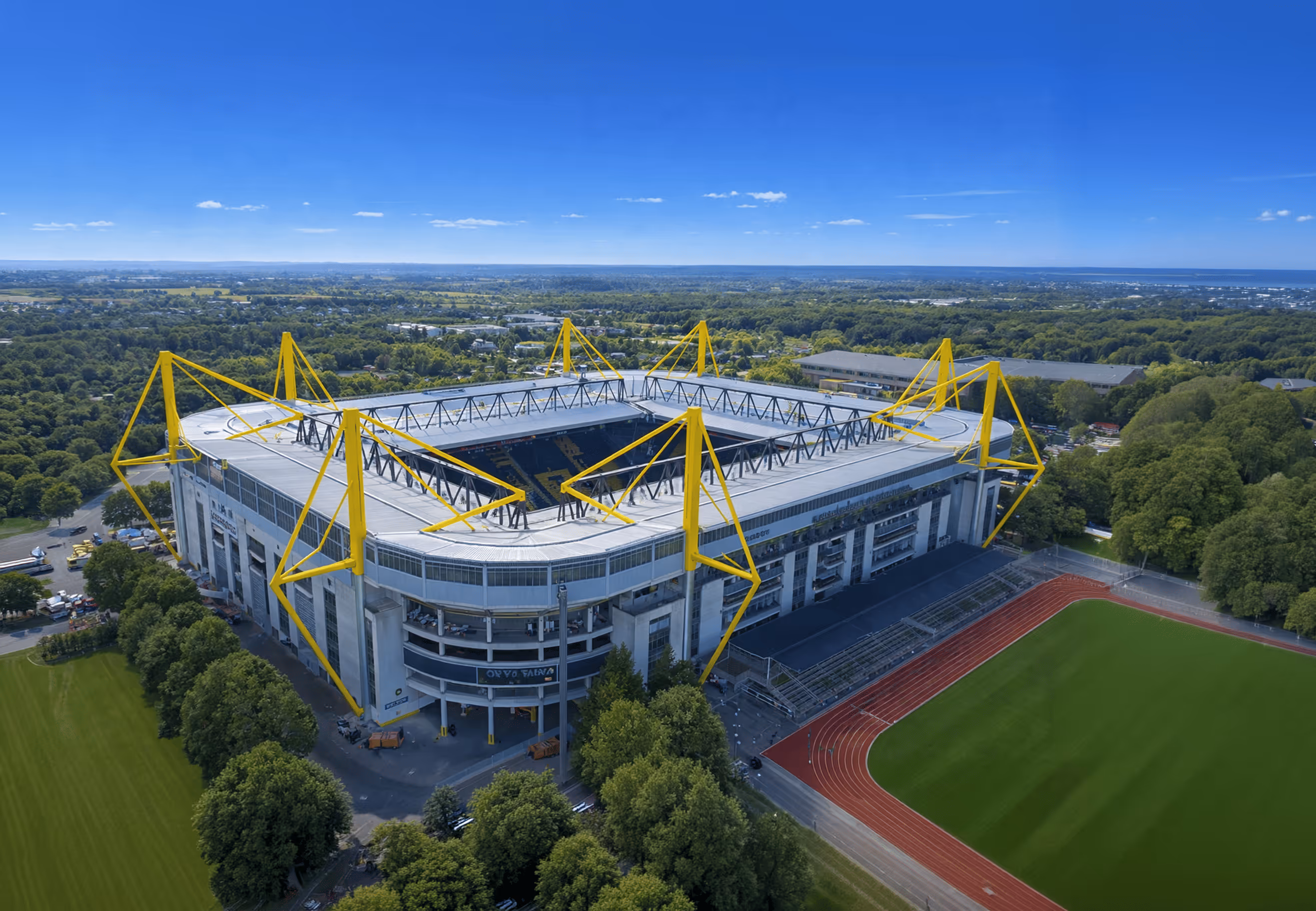 Luftaufnahme eines großen Stadions mit markanten gelben Stützstreben und einer Laufbahn um das Spielfeld, umgeben von grünen Bäumen.