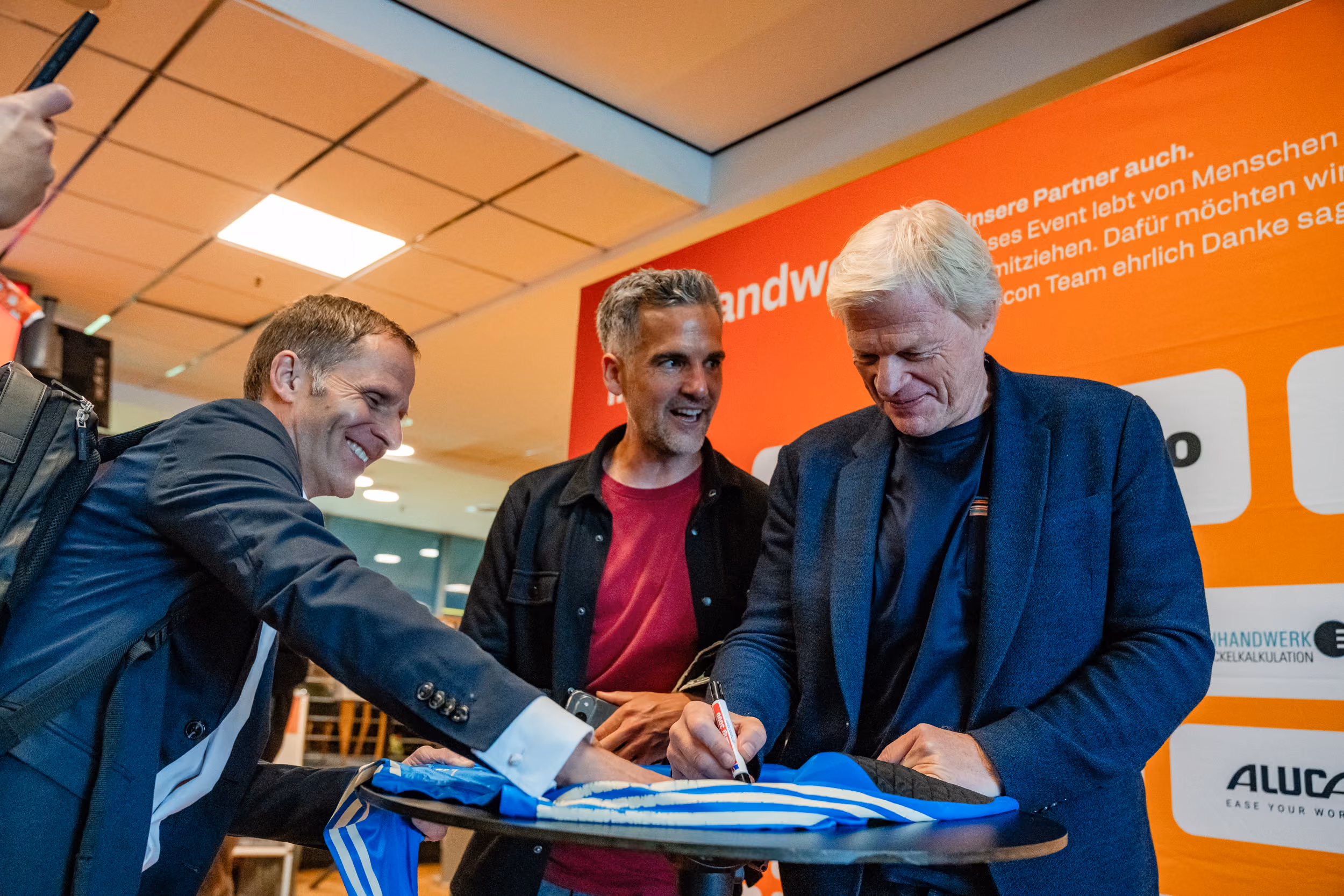 Drei Männer in einem Innenraum, einer unterschreibt ein blau-weiß gestreiftes Trikot auf einem erhöhten runden Tisch, während die anderen lächeln und zuschauen, mit orangefarbenem Präsentationshintergrund.