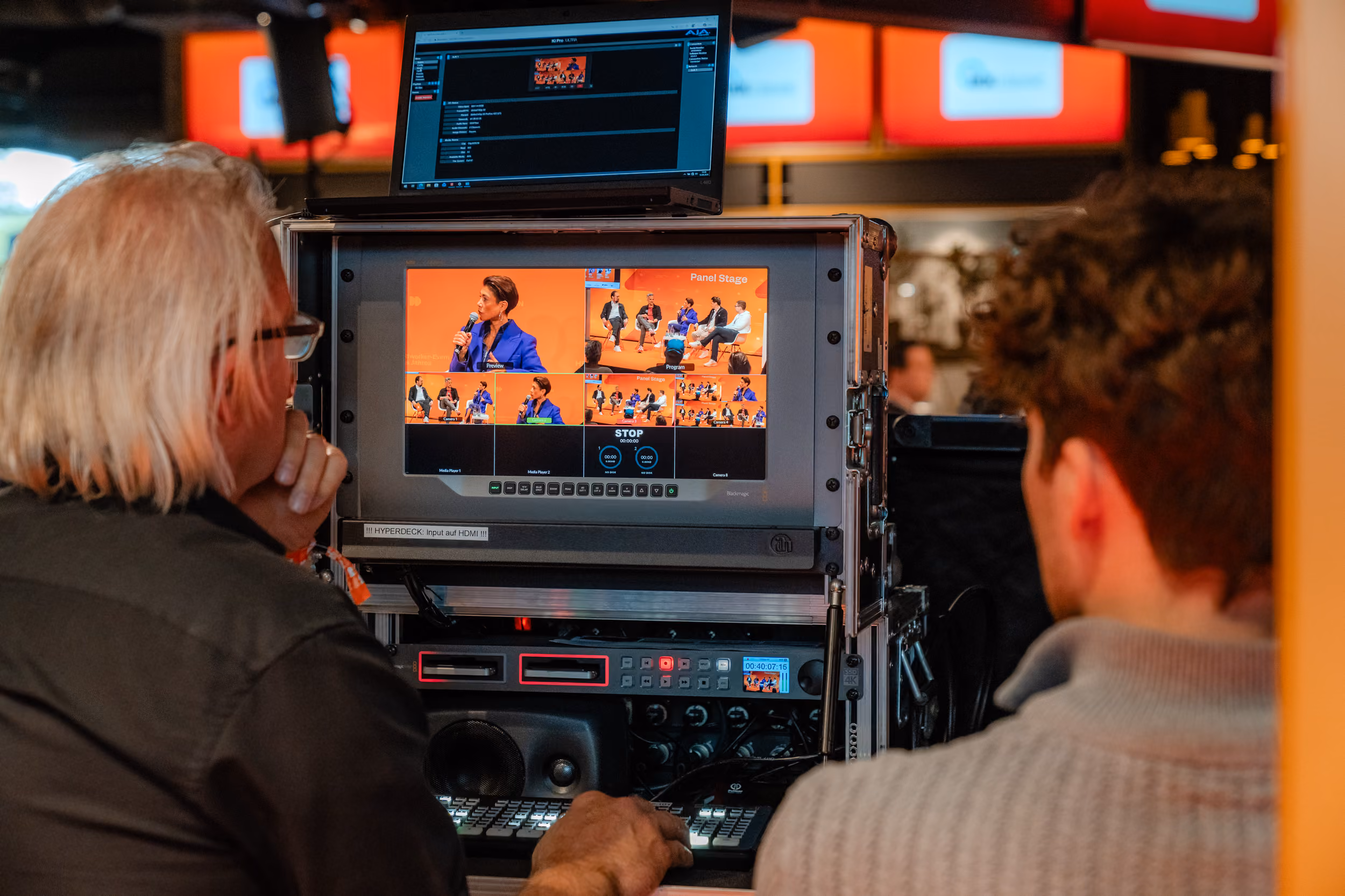 Zwei Personen beobachten einen Bildschirm mit mehreren Videoansichten eines Podiumsgesprächs, bei dem sechs Personen auf einer Bühne sitzen, eine Frau spricht mit einem Mikrofon.