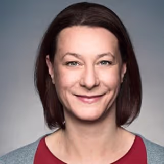 Studio-Porträt einer Frau mit schulterlangen braunen Haaren, rotem Oberteil und freundlichem Gesichtsausdruck vor grauem Hintergrund.