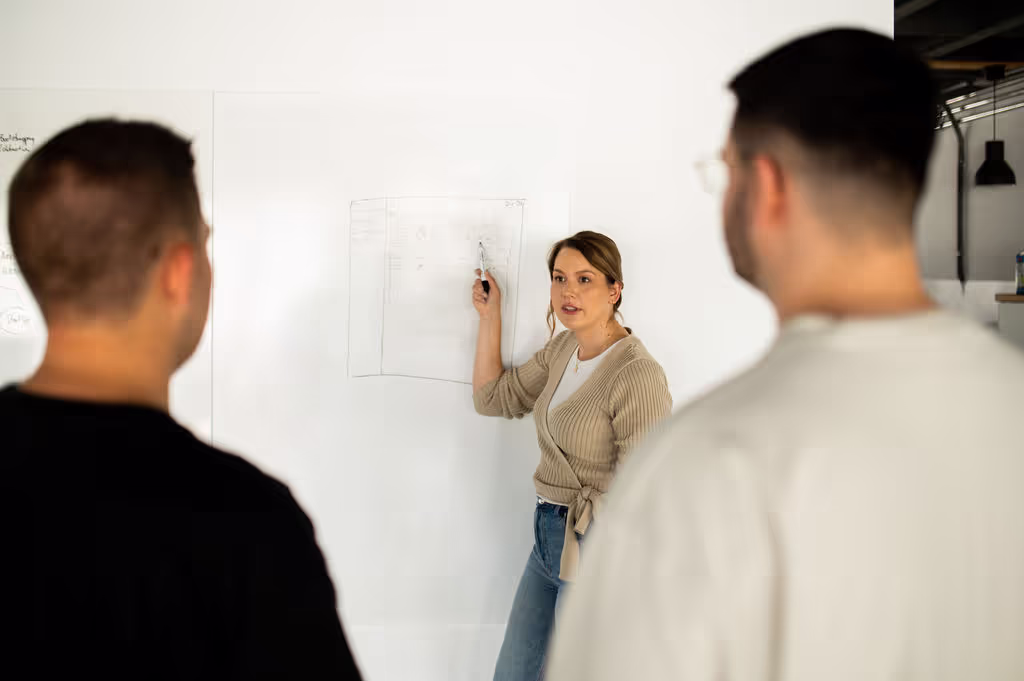 Frau präsentiert vor einem Whiteboard, zwei Männer hören zu. Szene aus einem UX-Workshop, Meeting oder interner Schulung.