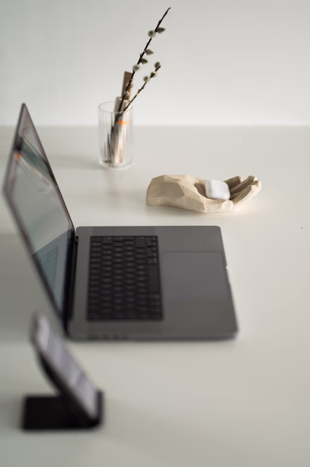 Heller Arbeitsplatz mit aufgeklapptem Laptop, Trockenblume und kleiner Skulptur auf einem Tisch. Reduziertes Setting für konzentriertes Arbeiten.
