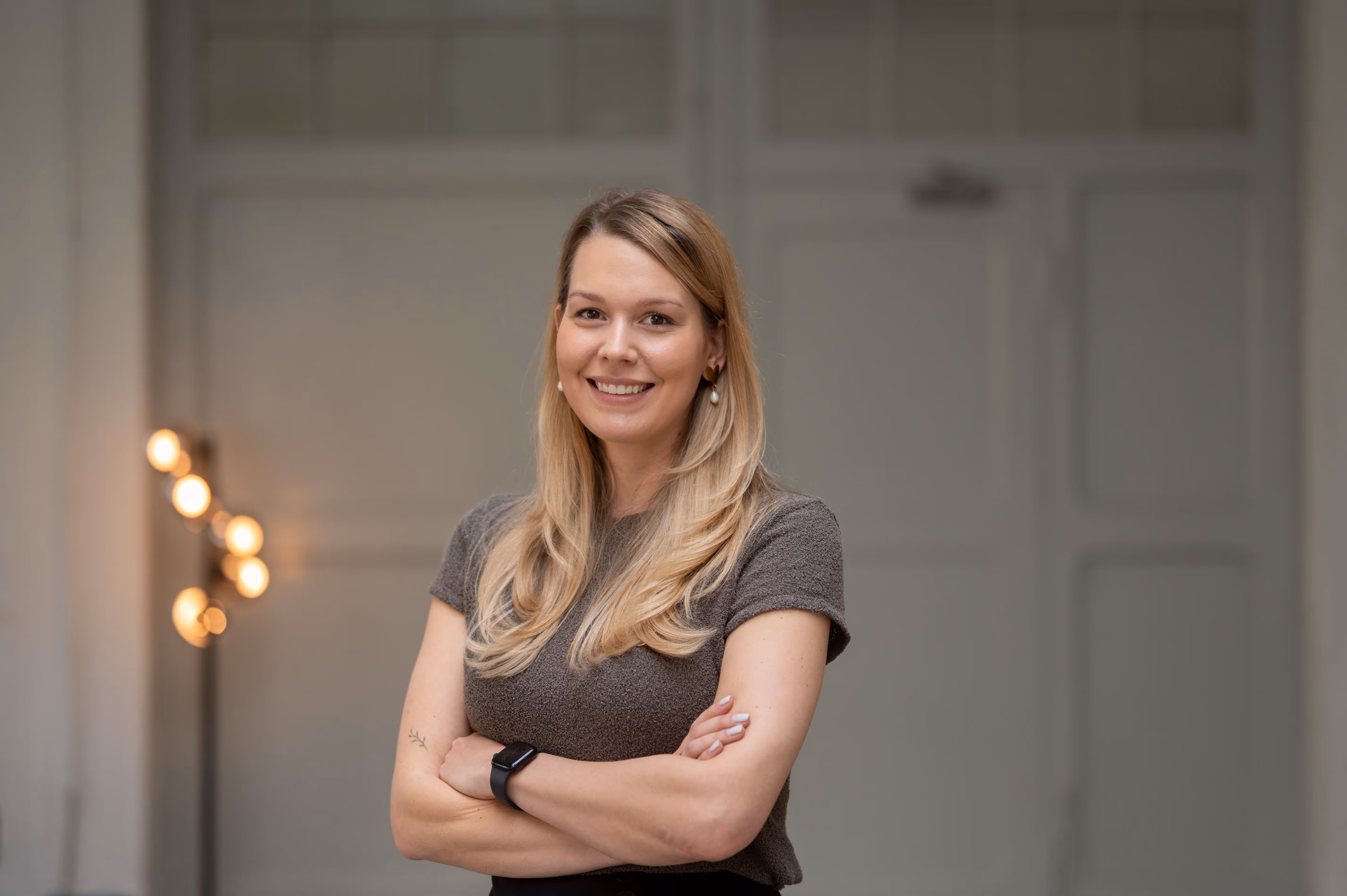 Portrait einer lächelnden Frau mit langen, blondem Haar in einem minimalistischen Innenraum mit weicher Beleuchtung. Sie steht mit verschränkten Armen vor einer weißen Doppeltür, trägt ein schlichtes Shirt und wirkt professionell und nahbar. Symbolbild für Leadership, UX-Expertise und modernes Unternehmertum.
