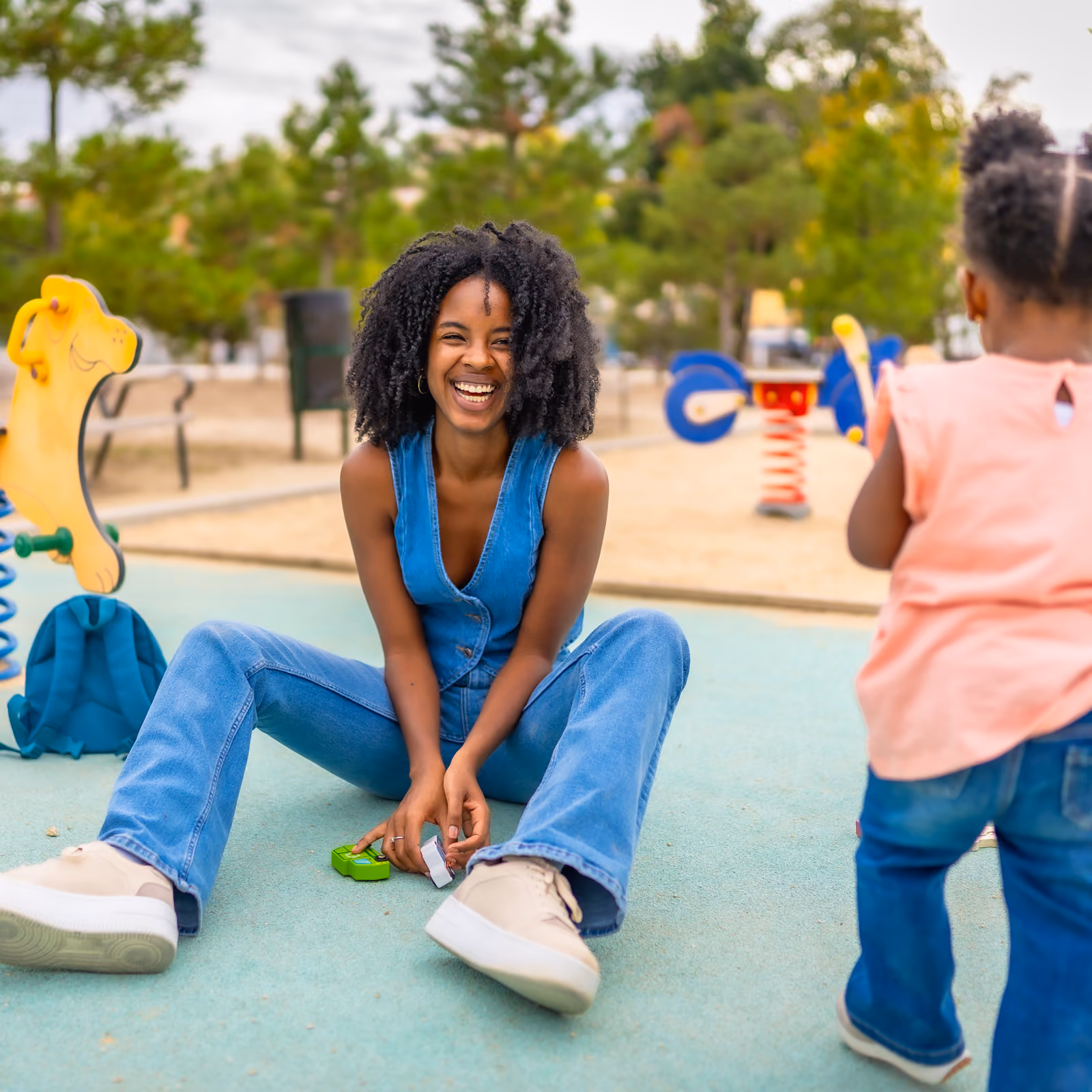Smiling woman in blue overalls sitting on playground floor playing with a child.