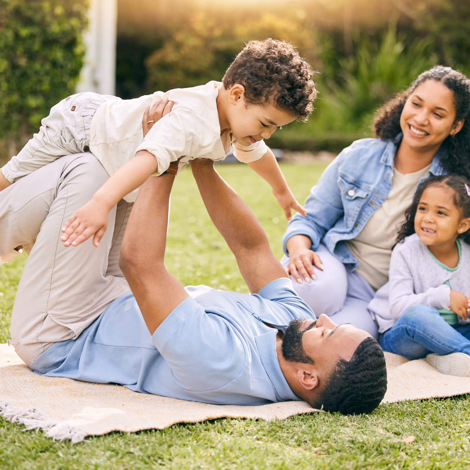 Father lying on blanket in yard holding son up while mother and daughter watch smiling.