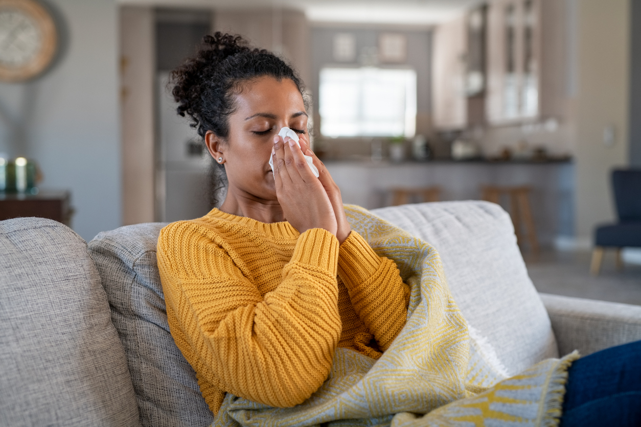 Person sneezing into a tissue while bundled in a sweater near a frosted window