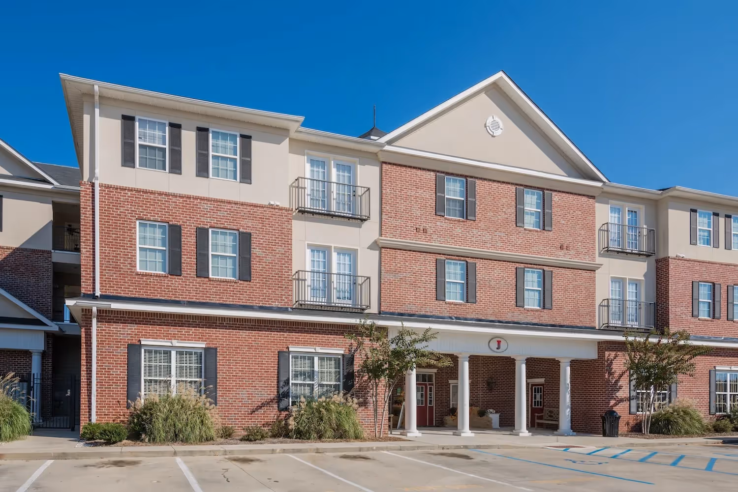 Exterior facade of The Grand at Lake Sheri apartment building in Valdosta, GA, showcasing brick and beige siding, with balconies and a covered entrance.