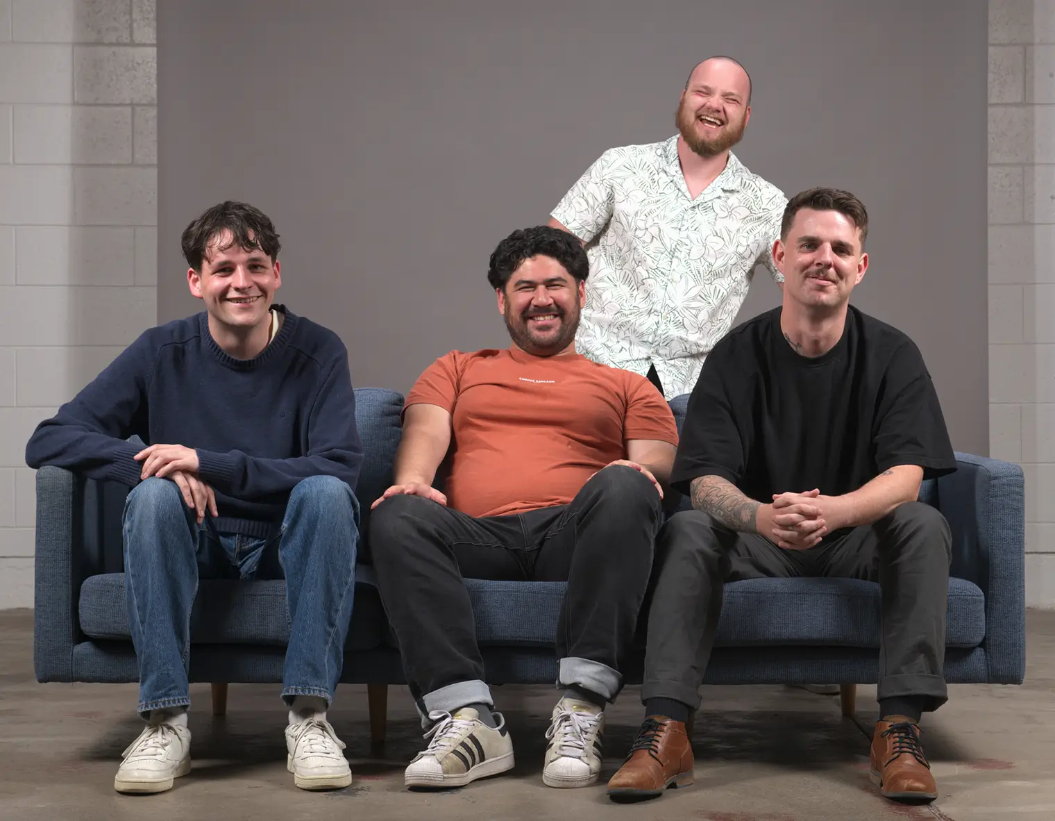 Four men smiling, three seated on a blue couch and one standing behind them against a gray background.