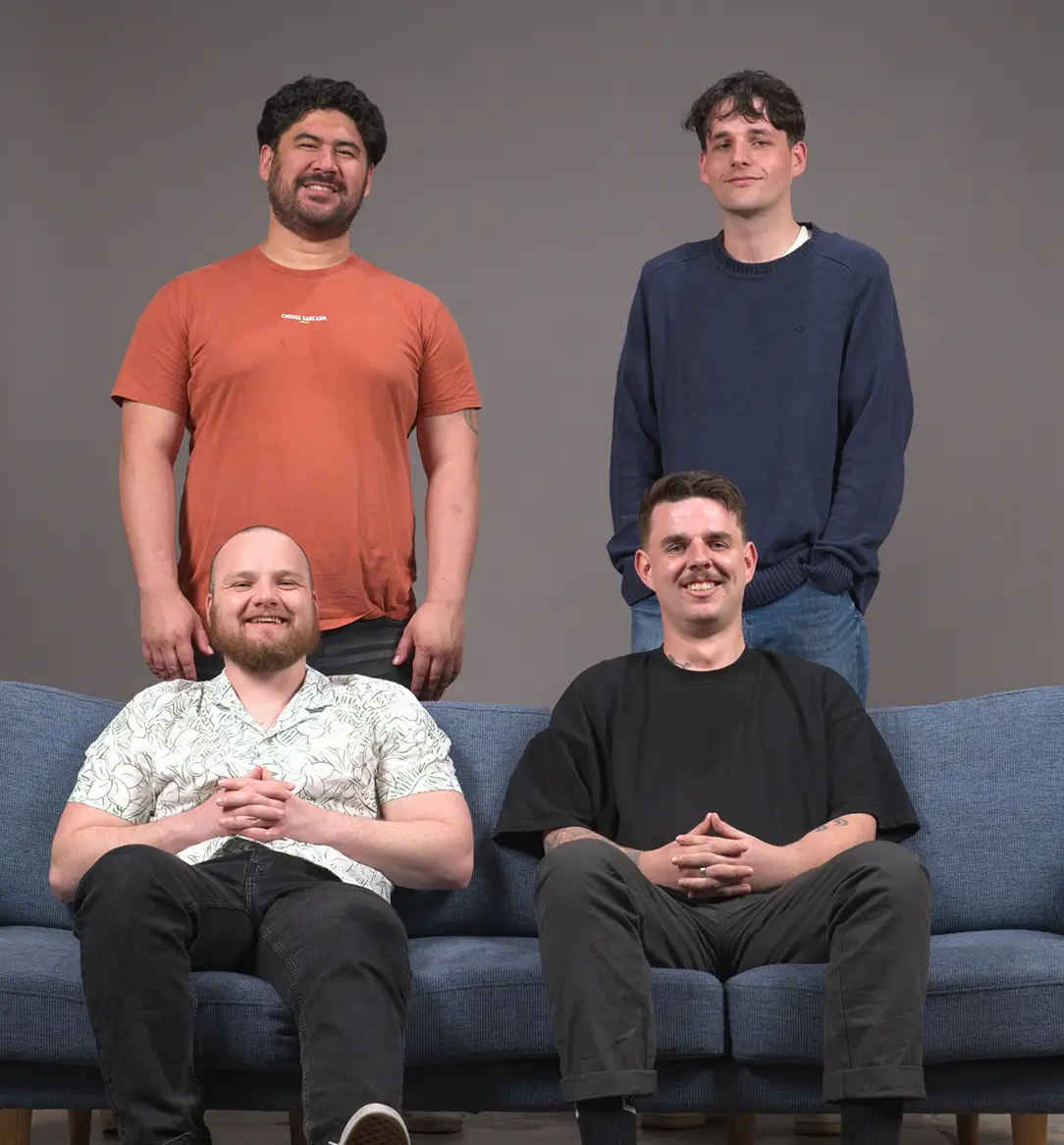 Four men smiling, two sitting on a blue couch and two standing behind them against a plain gray background.