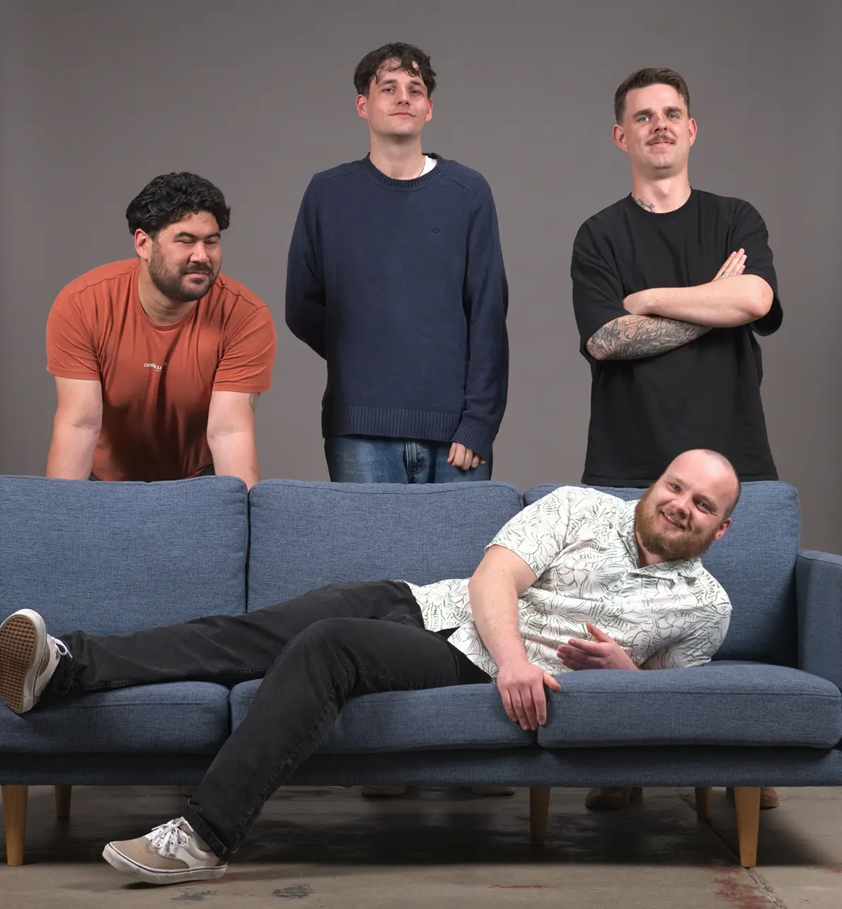 Four men posing with three standing behind a blue couch and one lying down on it, all smiling or relaxed.