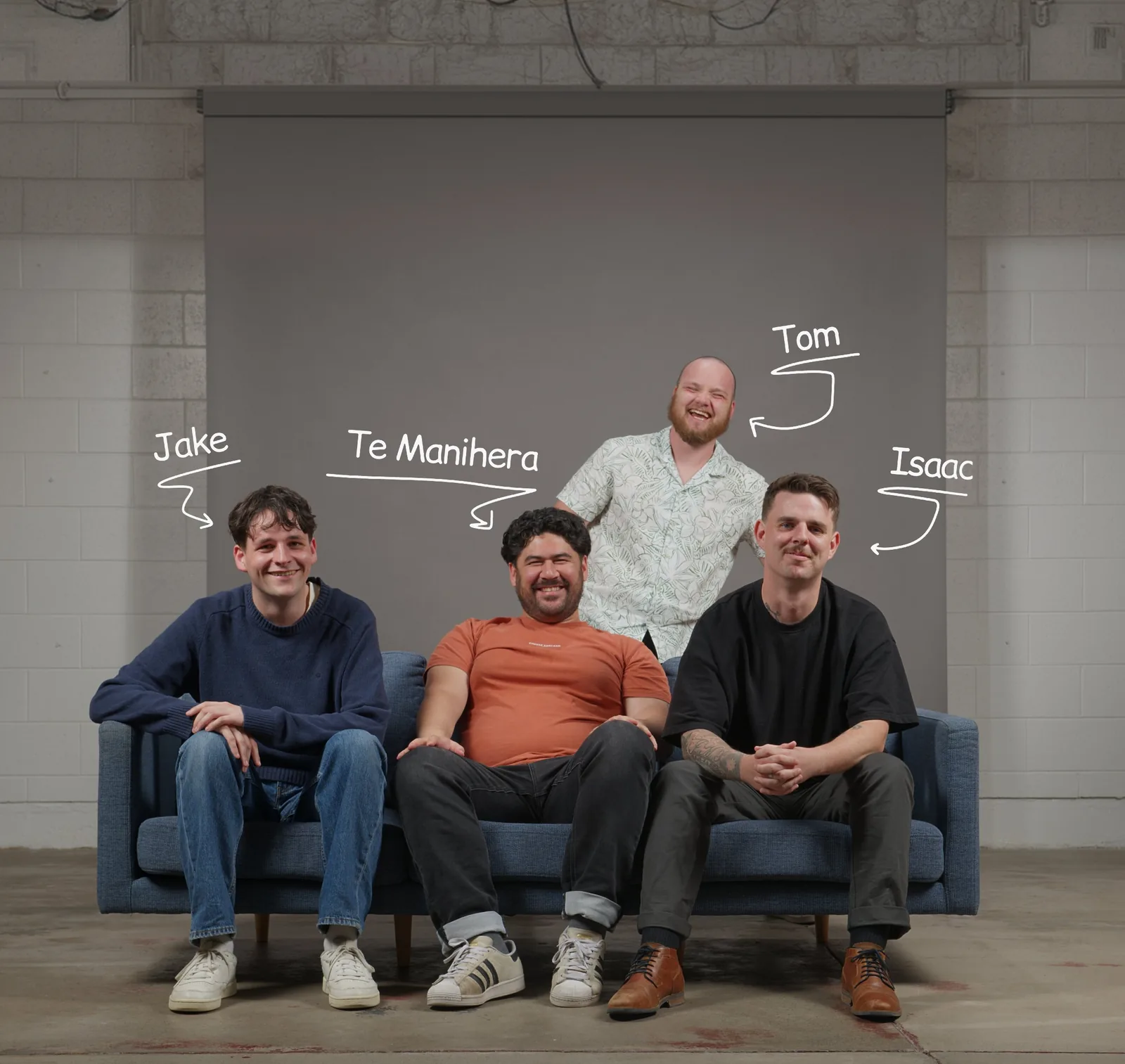 Four men posing for a photo in a room with concrete walls and floor, three seated on a blue couch and one standing behind. From left to right on the couch are Jake in a blue sweater and jeans, Te Manihera in an orange t-shirt and dark pants, and Isaac in a black t-shirt with tattoos on his right forearm wearing brown shoes. Standing behind the couch is Tom, smiling broadly in a light-patterned shirt. Each man has his name written above with an arrow pointing to him.