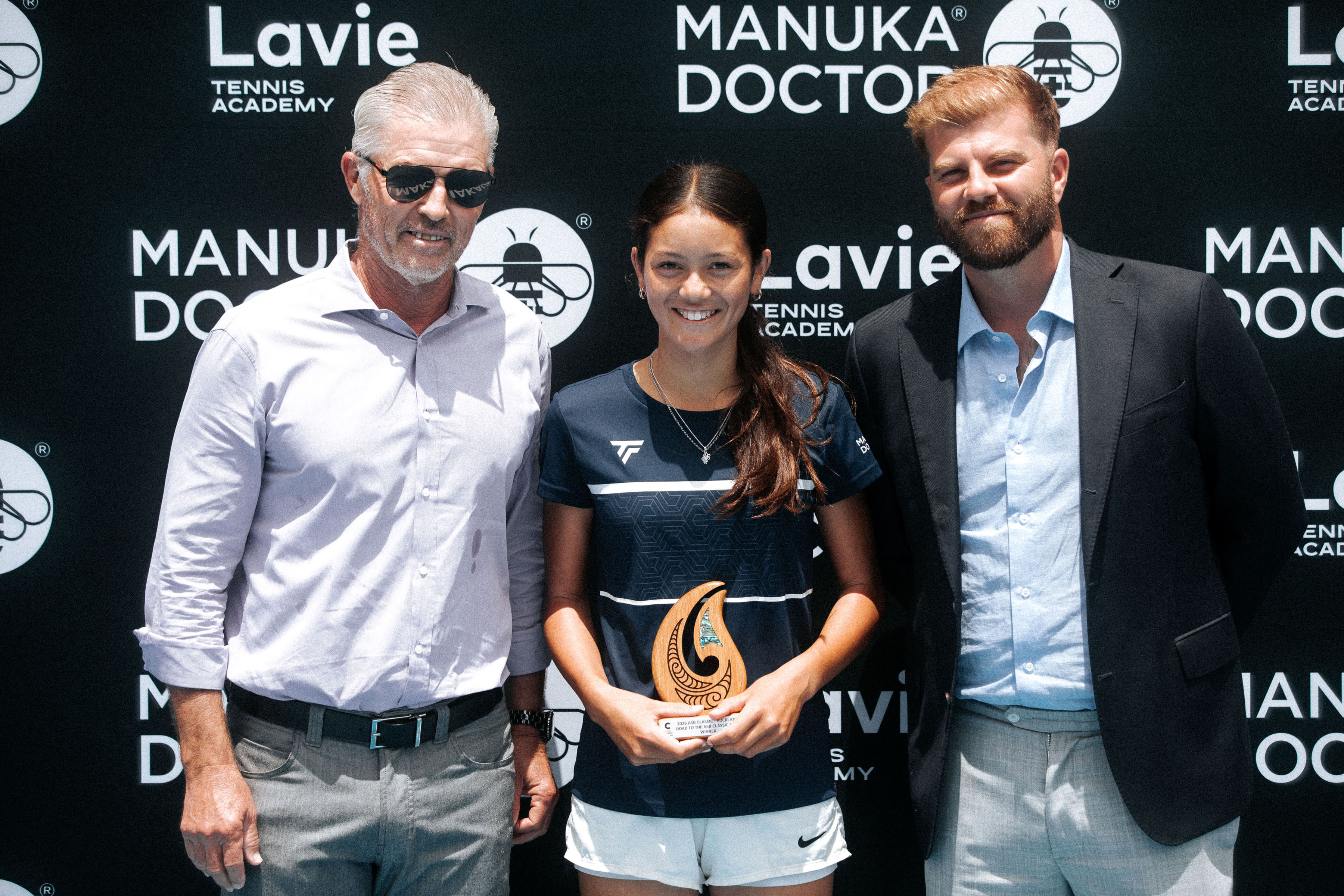 Road to the ASB Classic girls winner with her trophy in front of the Manuka Doctor and Lavie Tennis Academy wall