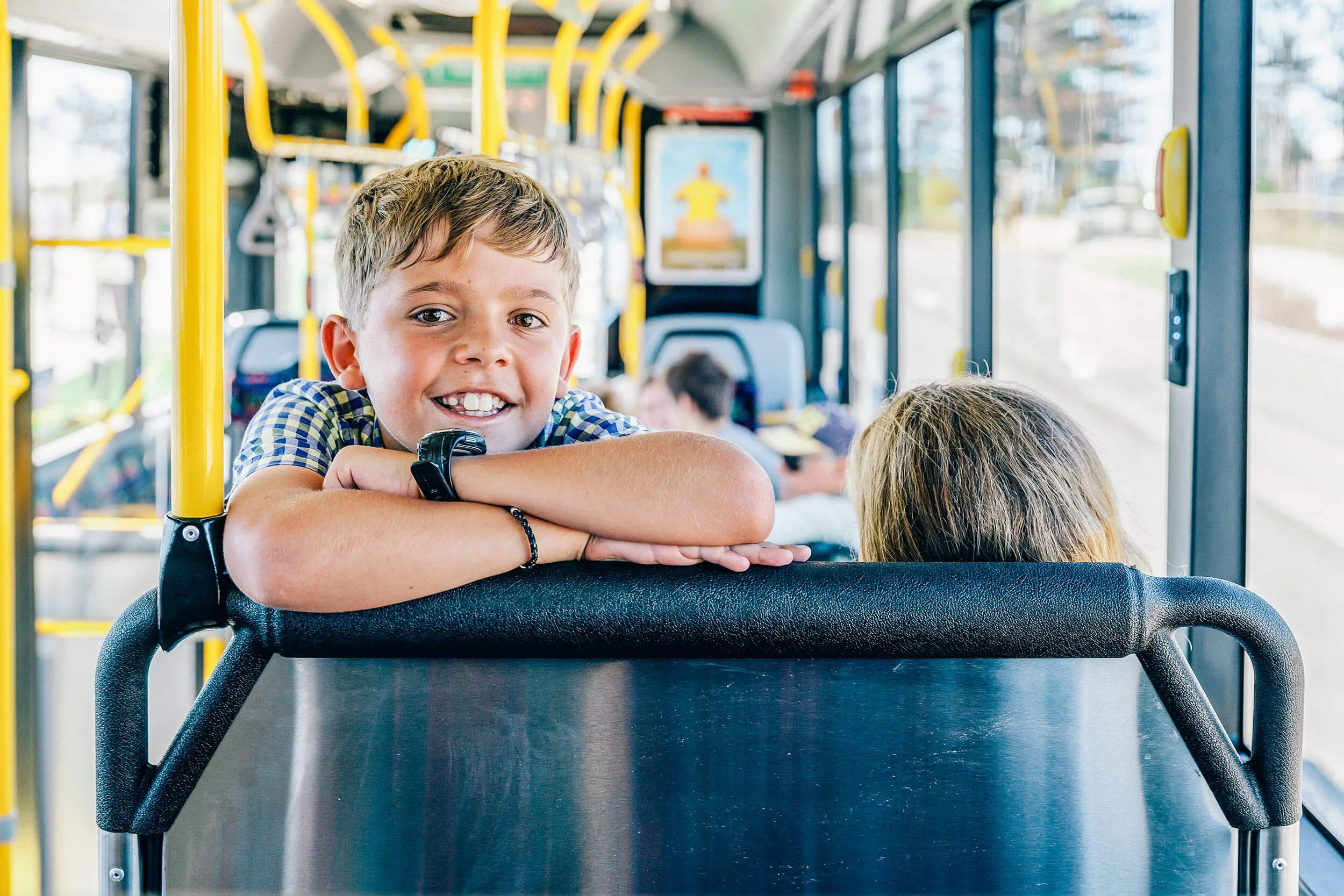 A young boy with a wide smile leaning on the back of a bus seat, looking directly at the camera.