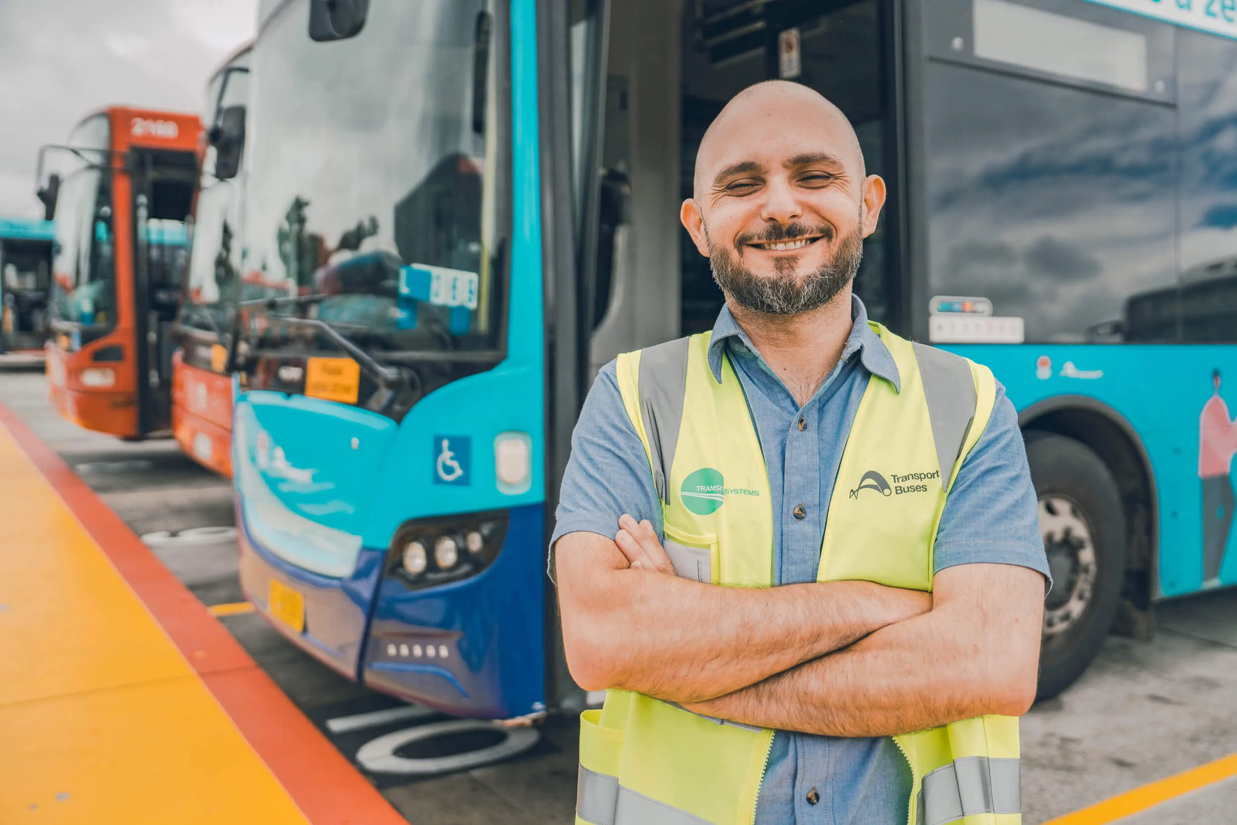 A smiling bald man with a beard, wearing a high-visibility vest with "Transport for Buses" logo, stands in front of buses.