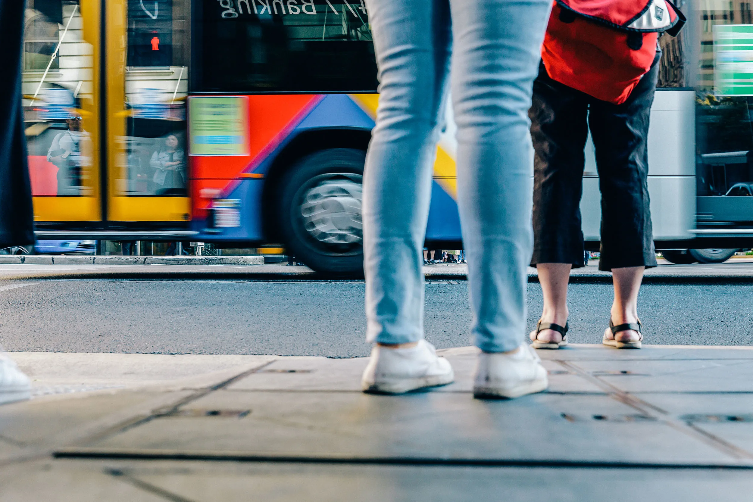 From a low angle, the lower legs of two people stand on a sidewalk, with a blurred colorful bus passing by in the background.