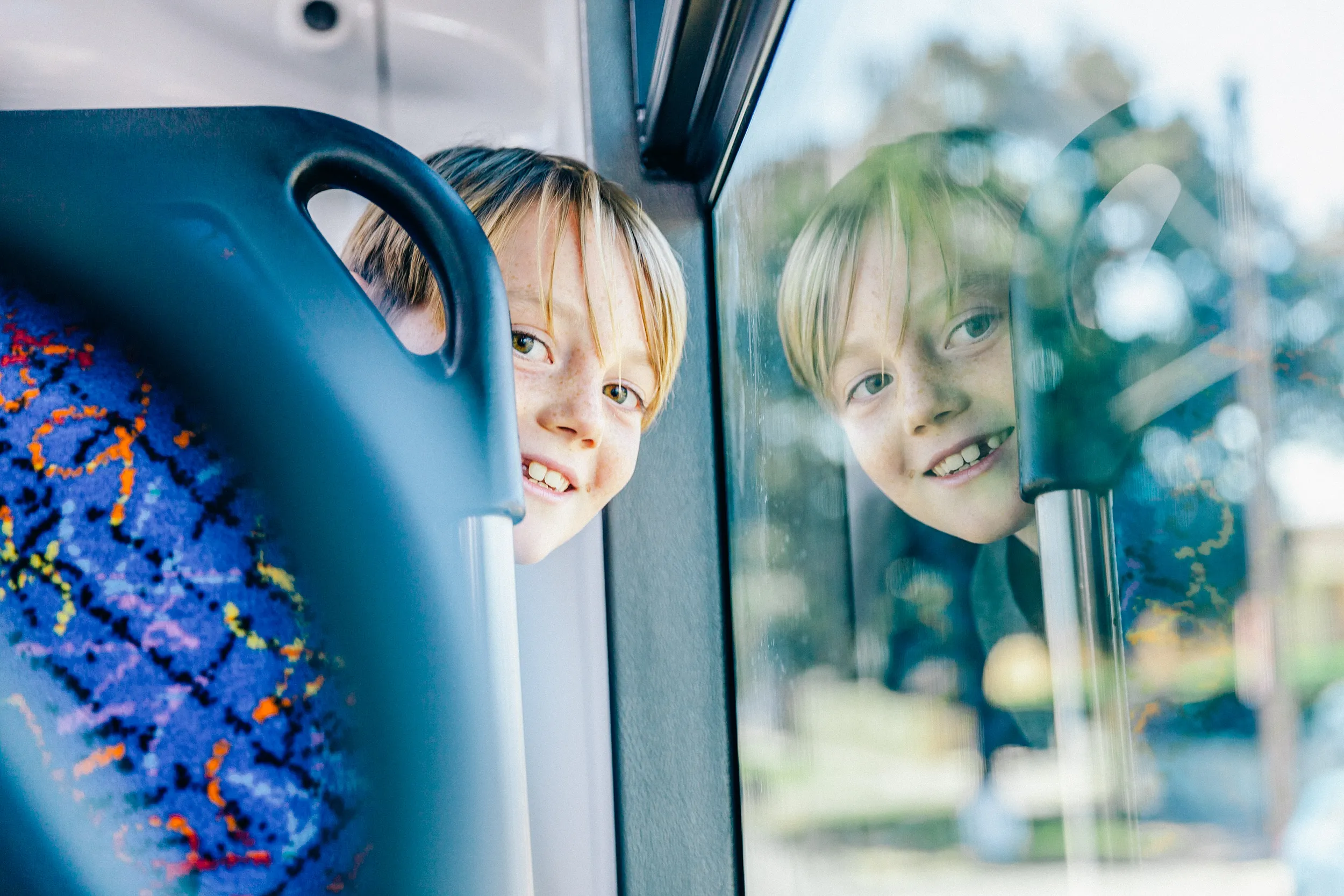 A smiling young boy with freckles looking out of a bus window, with his reflection visible.