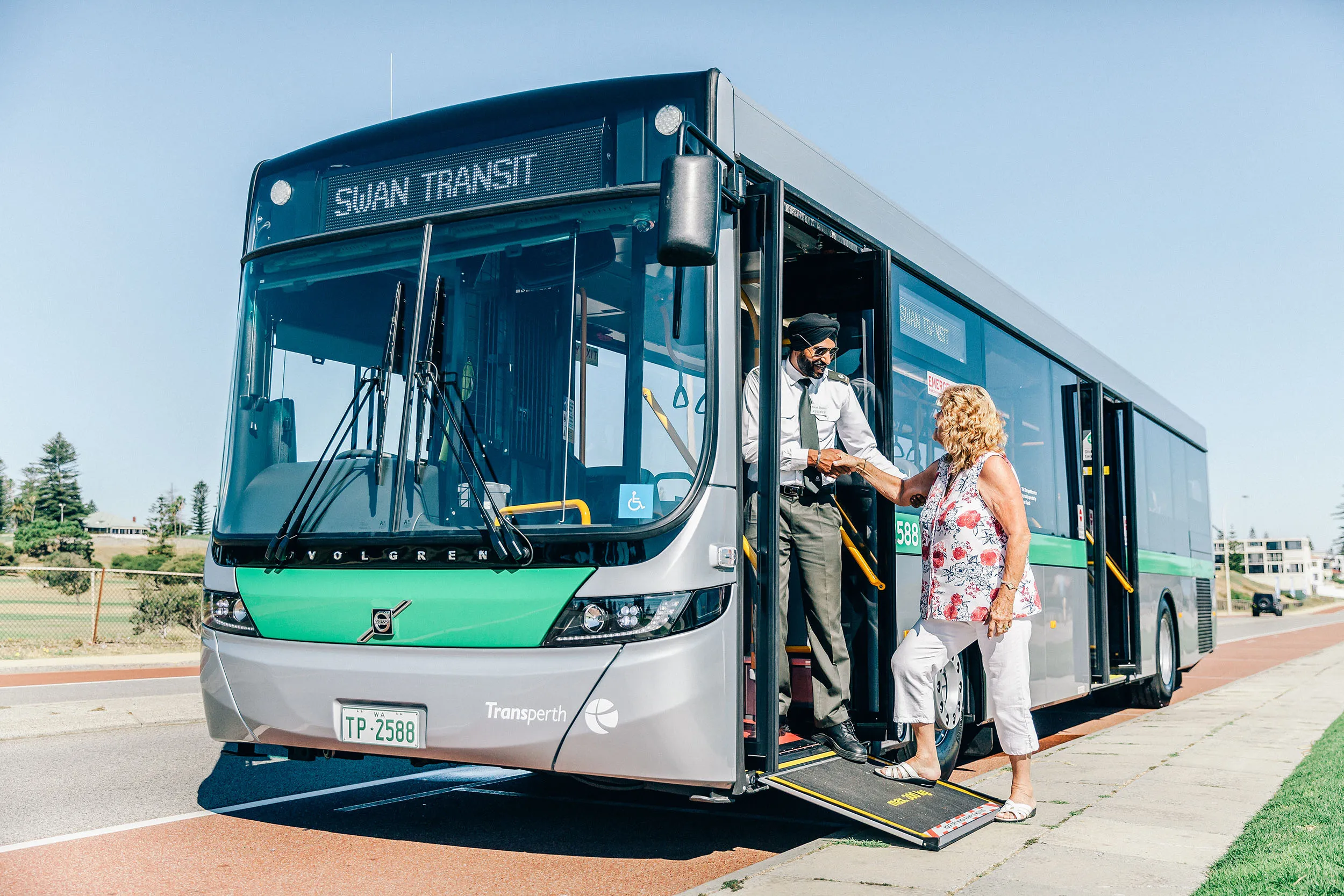 A male bus driver in a turban helping an elderly woman board a "SWAN TRANSIT Transperth" bus using a ramp.