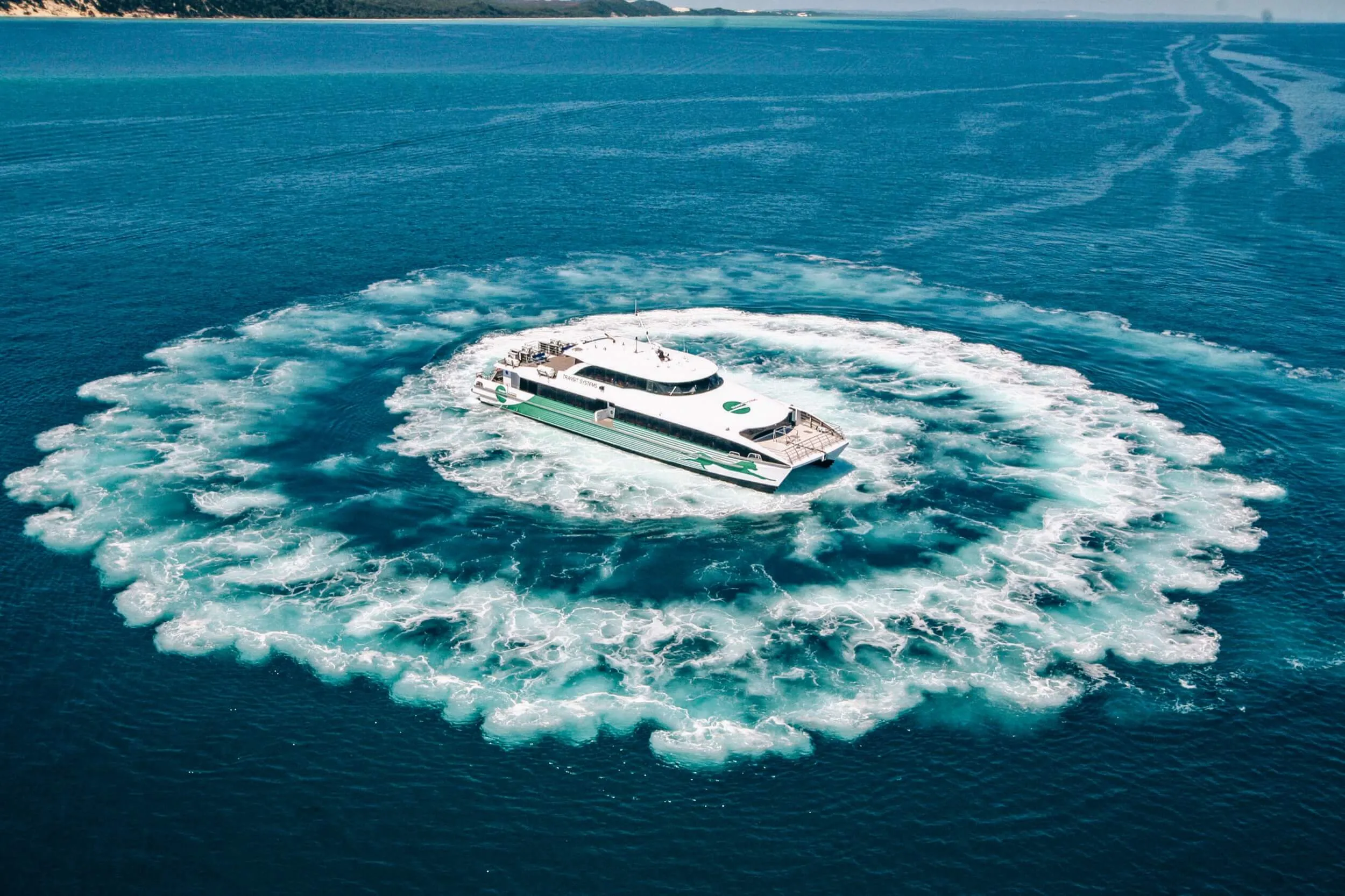 Aerial view of a white and green ferry making a circular wake in the clear blue ocean.