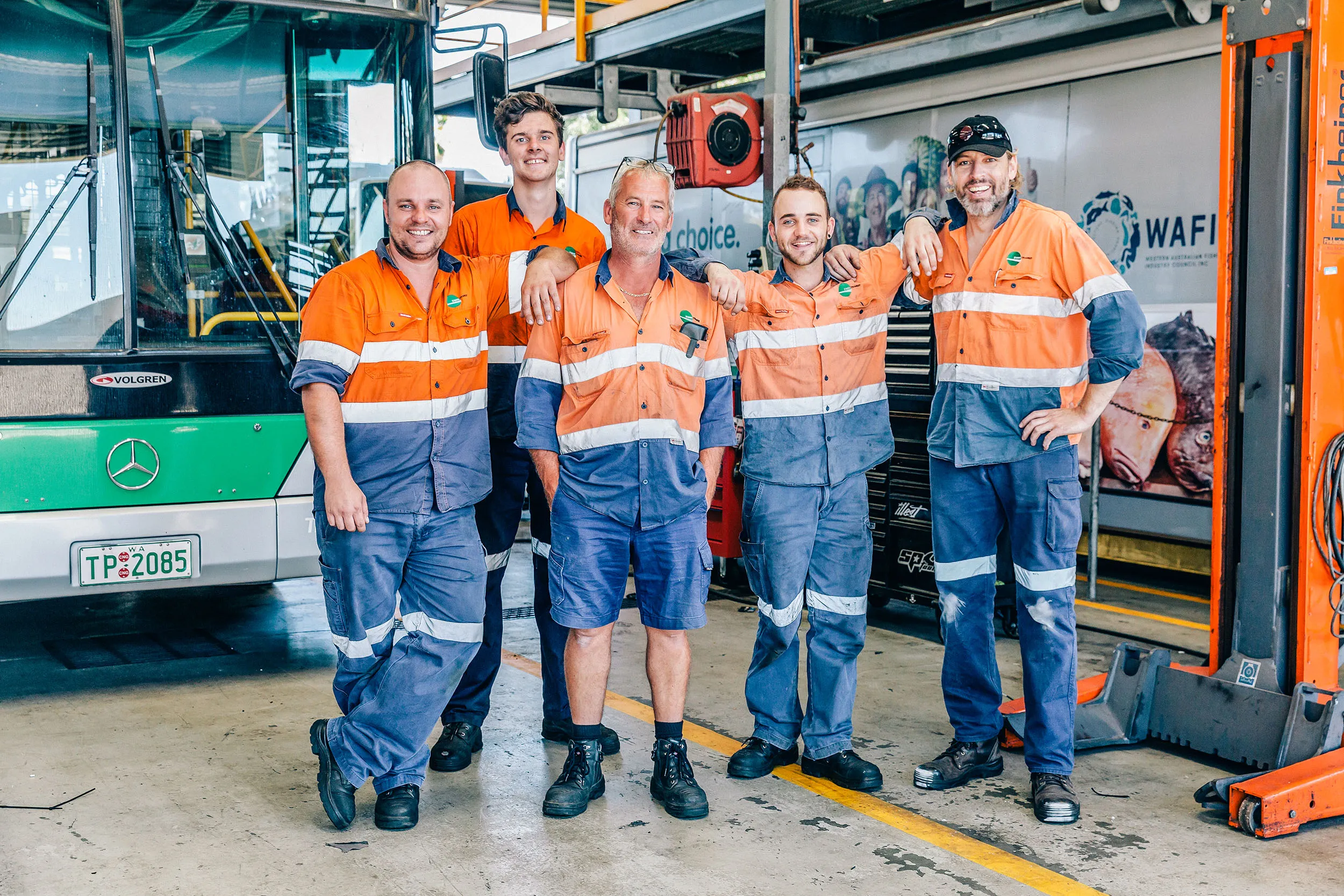 Five male mechanics in orange and blue work uniforms posing in front of a bus in a garage.